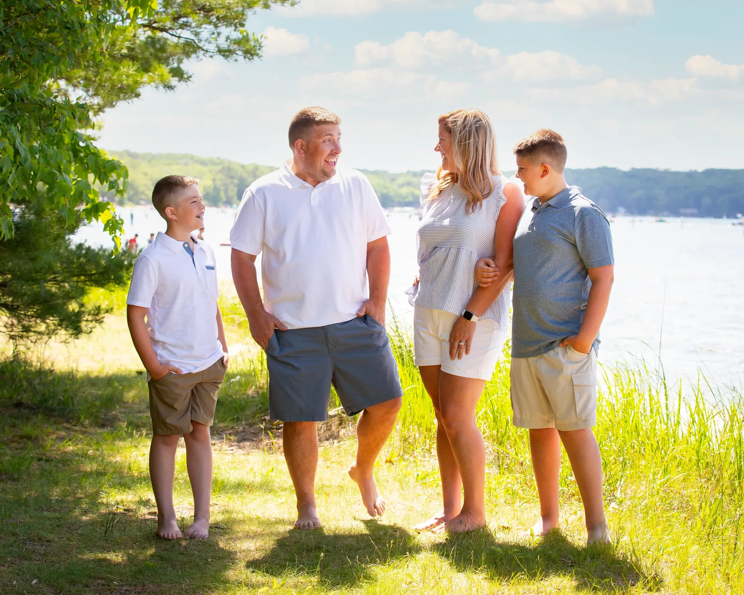 Family of four standing on grass near a lake, smiling and laughing together, with boats and trees in the background.