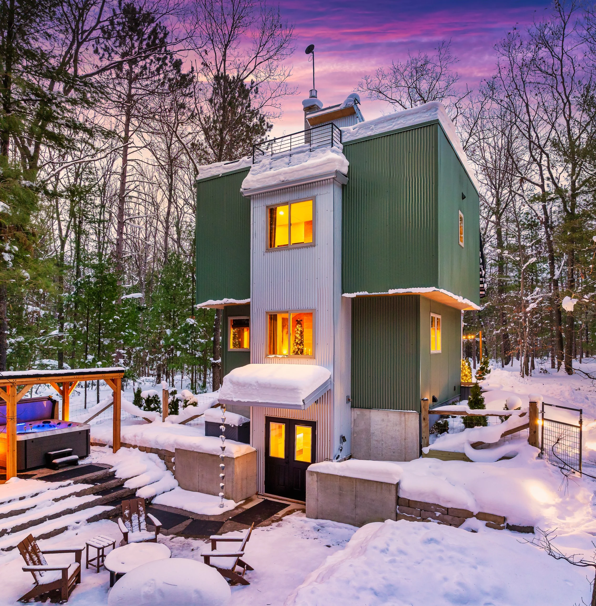 A modern multi-story house with green and white exterior, snow-covered roof and yard, illuminated windows, and surrounded by leafless trees at sunset or dusk.