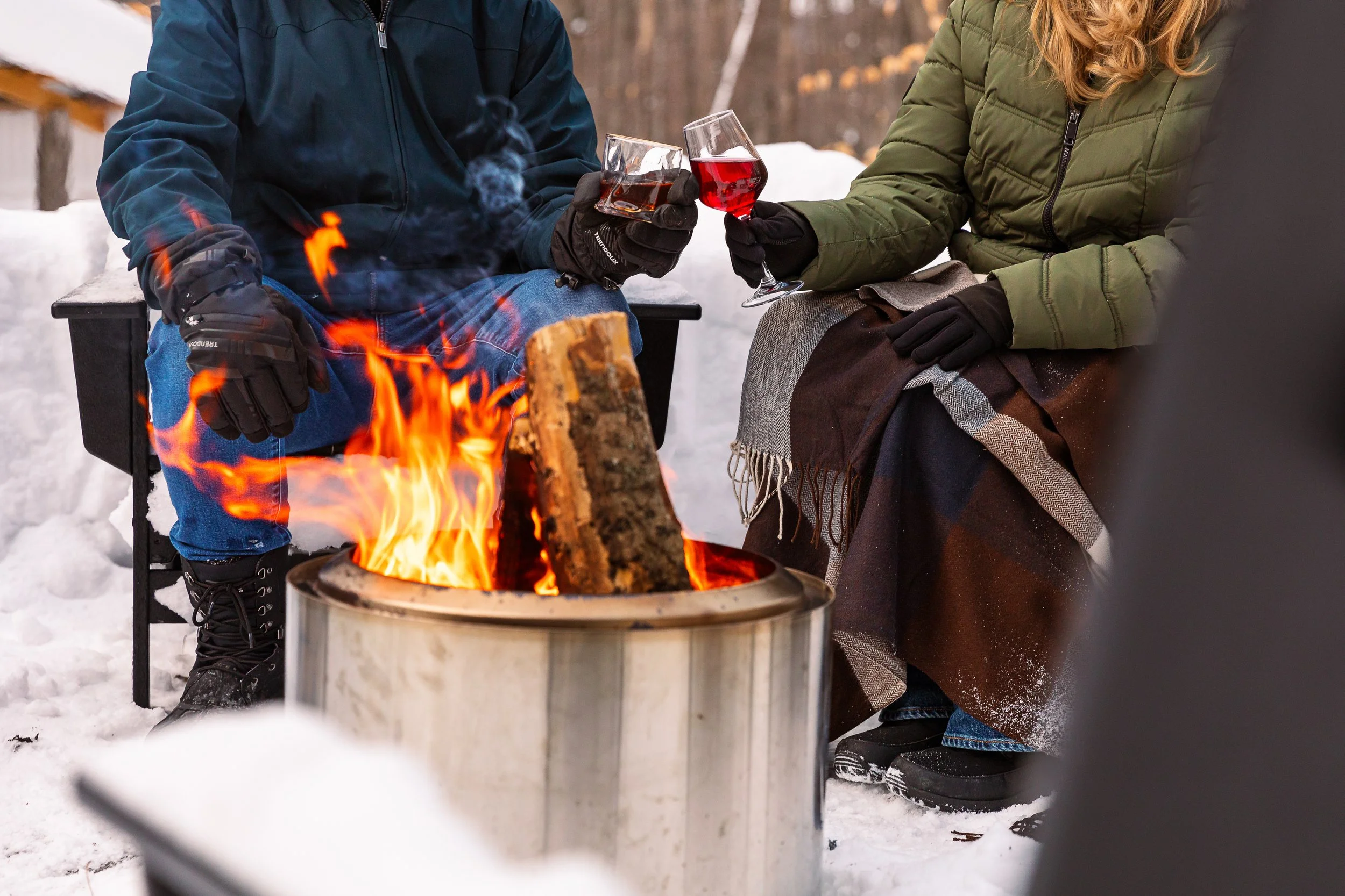 Two people sitting outside in winter clothes, warming by a campfire on snow, toasting glasses of red wine.
