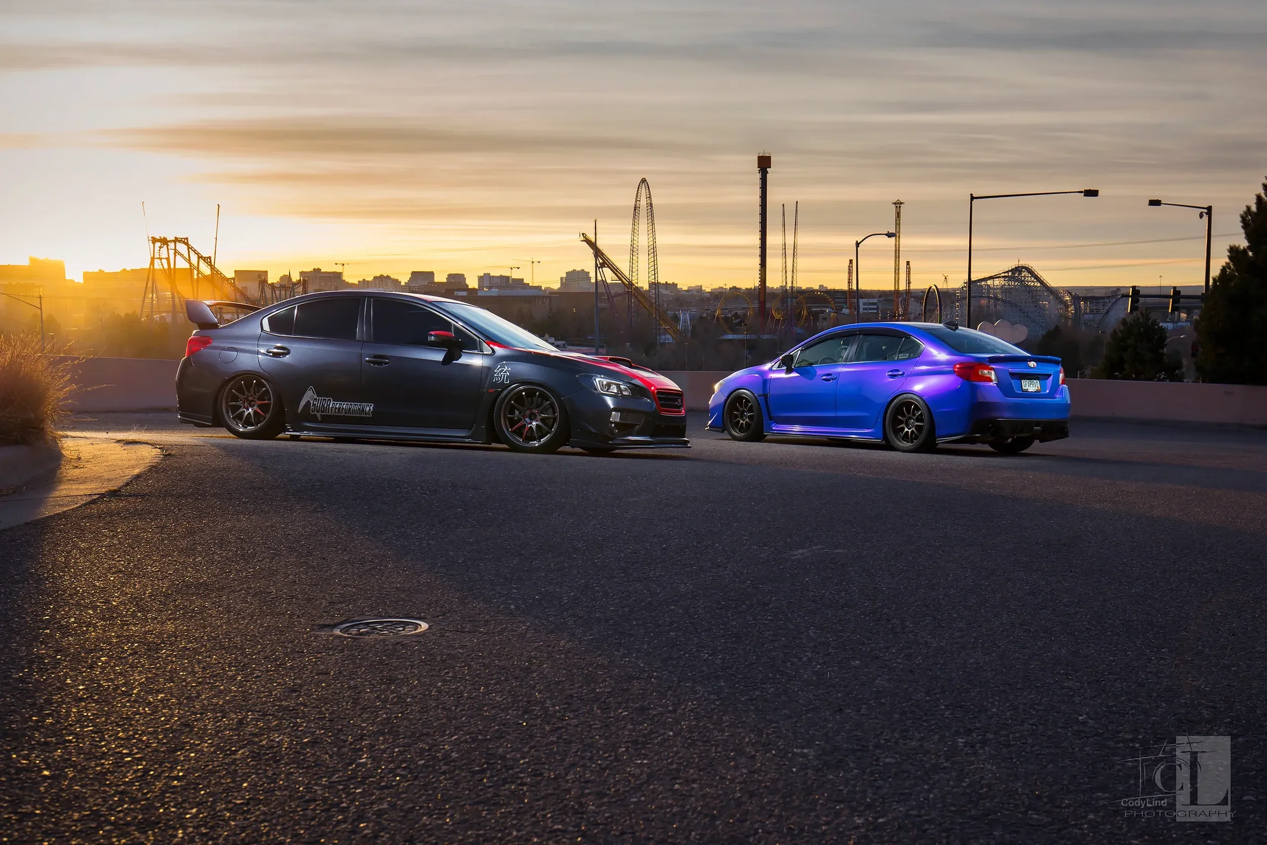 Two modified cars parked on a city street at sunset with an amusement park in the background.
