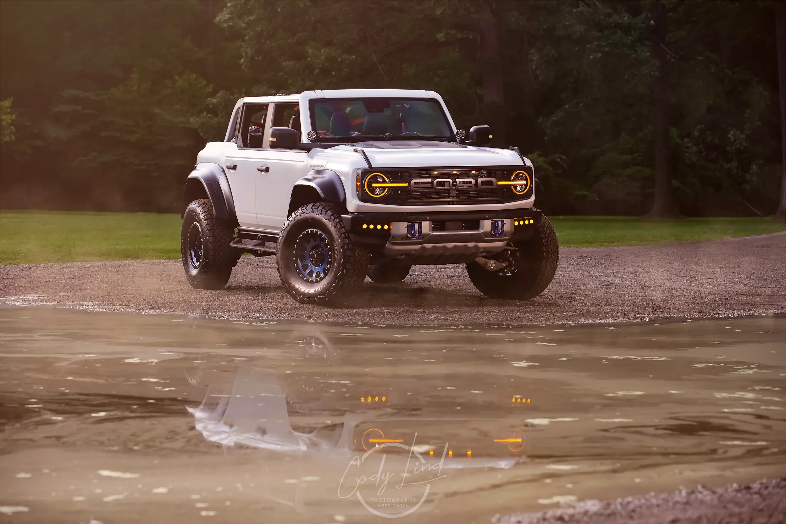 A white off-road truck with large tires and black fenders parked by a body of water on a gravel surface, with a backdrop of green trees.
