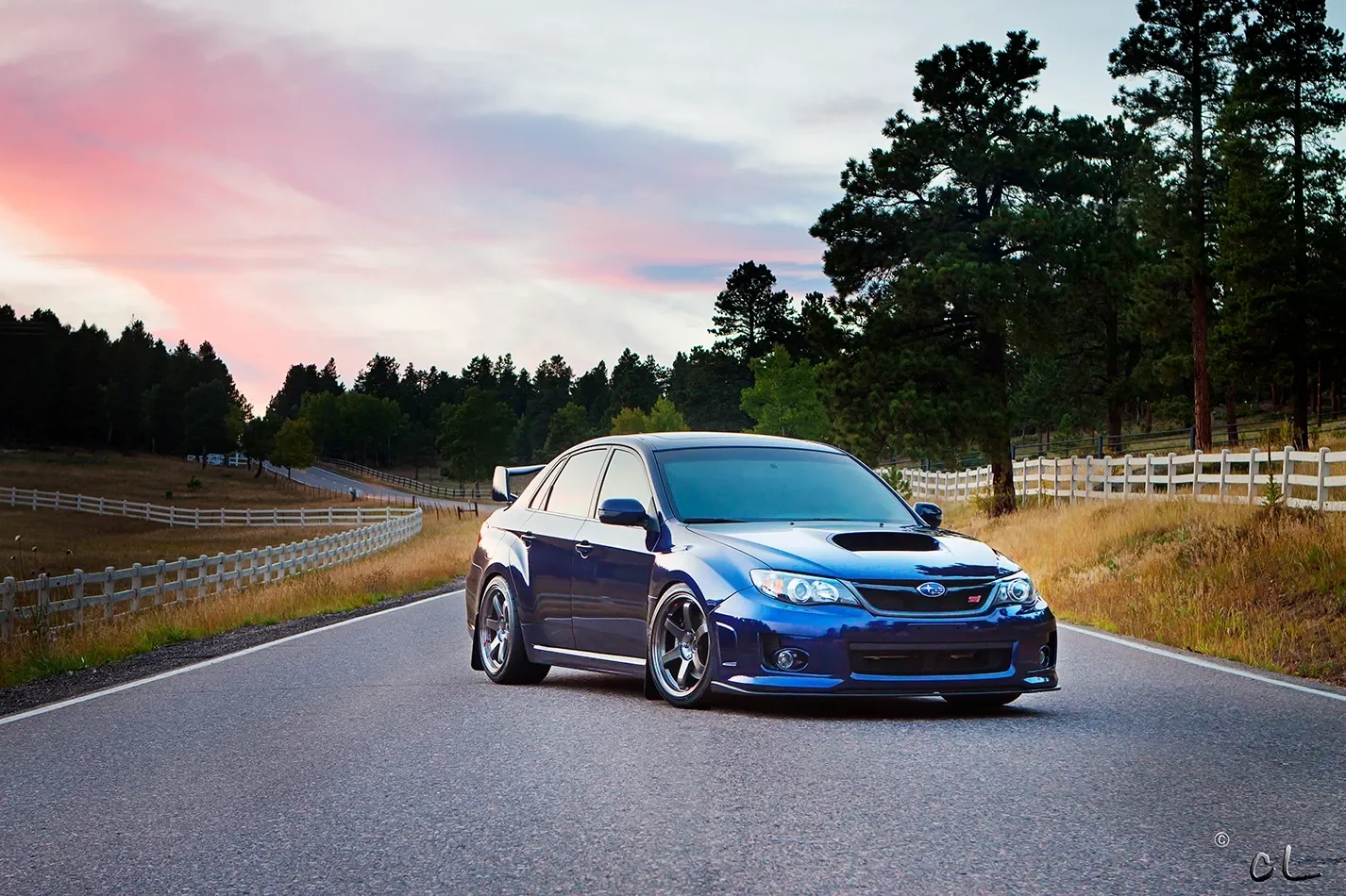A blue Subaru WRX car parked on a curved rural road during sunset, surrounded by tall trees and wooden fences.