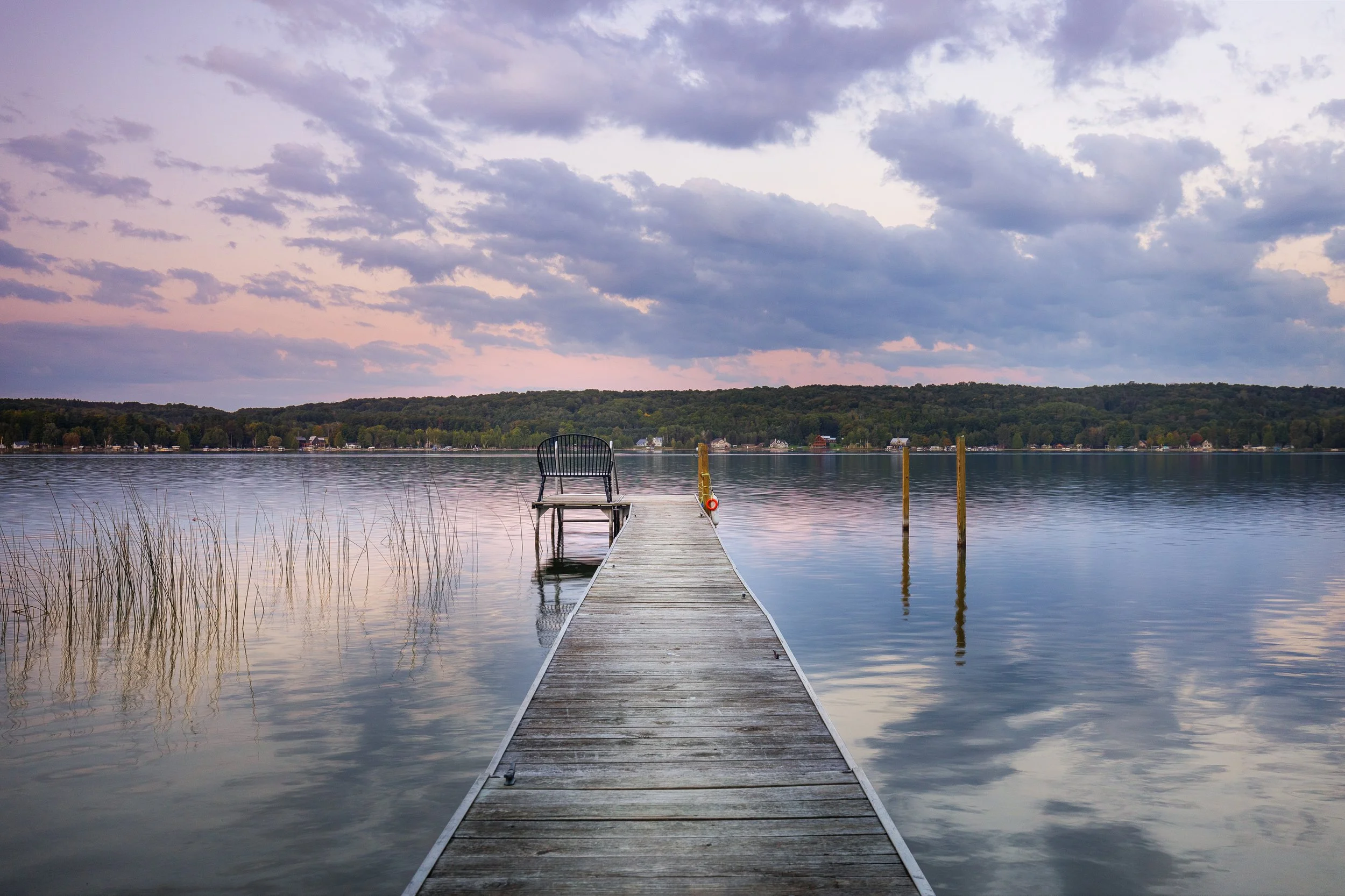 A wooden dock extends into a calm lake with a chair at the end, surrounded by still water and a cloudy sky overhead.