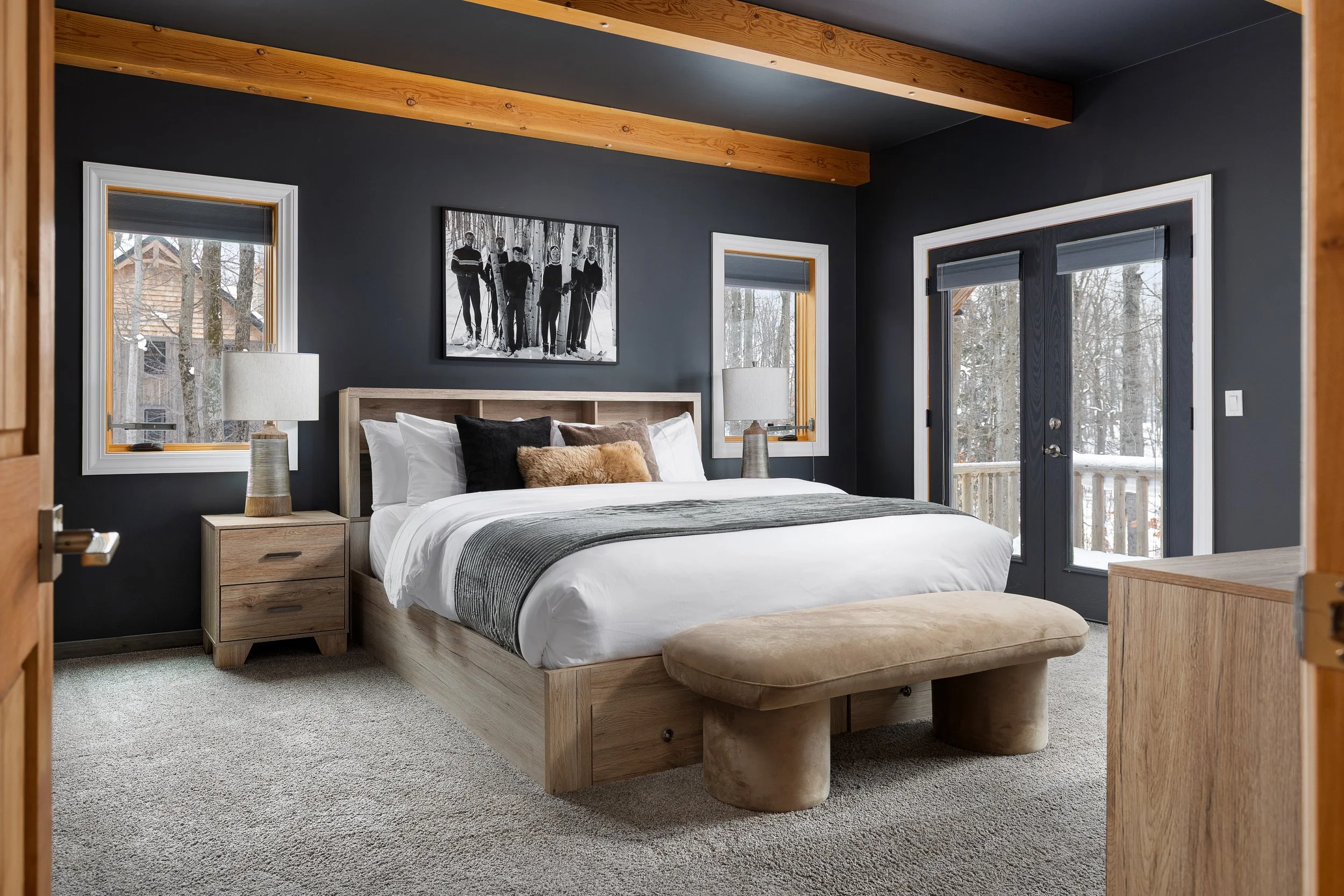 A bedroom with dark gray walls and wooden beams on the ceiling. It has two windows, a glass door leading outside, a wooden bed frame with white bedding and pillows, two bedside tables with lamps, a wall hanging photo of people in ski gear, and an uph