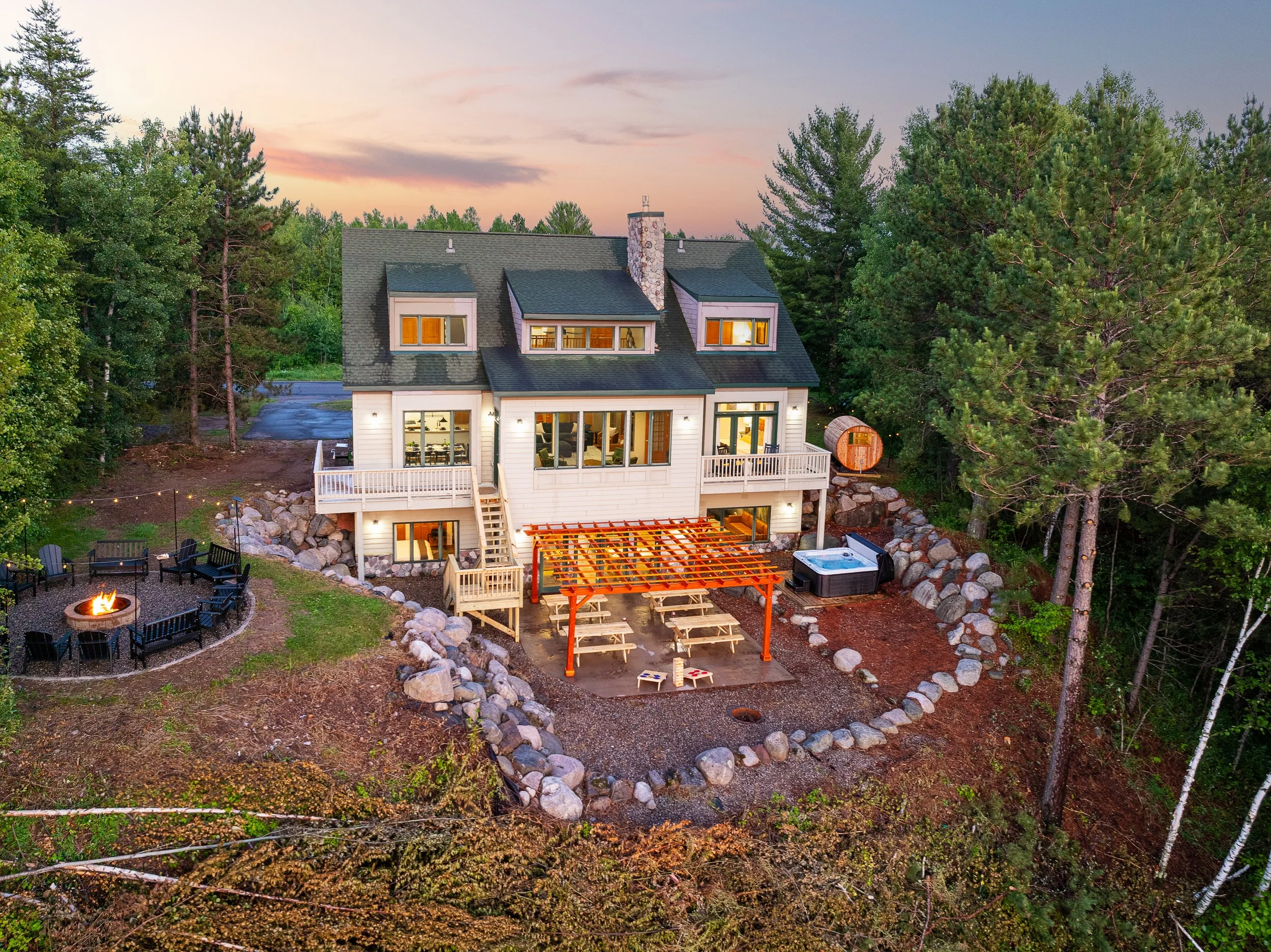 A multi-story house with a grey shingled roof and white siding, surrounded by tall green trees, during sunset. The backyard features a fire pit with black chairs, a hot tub, a small wooden barrel, and a patio with a pergola and picnic tables.