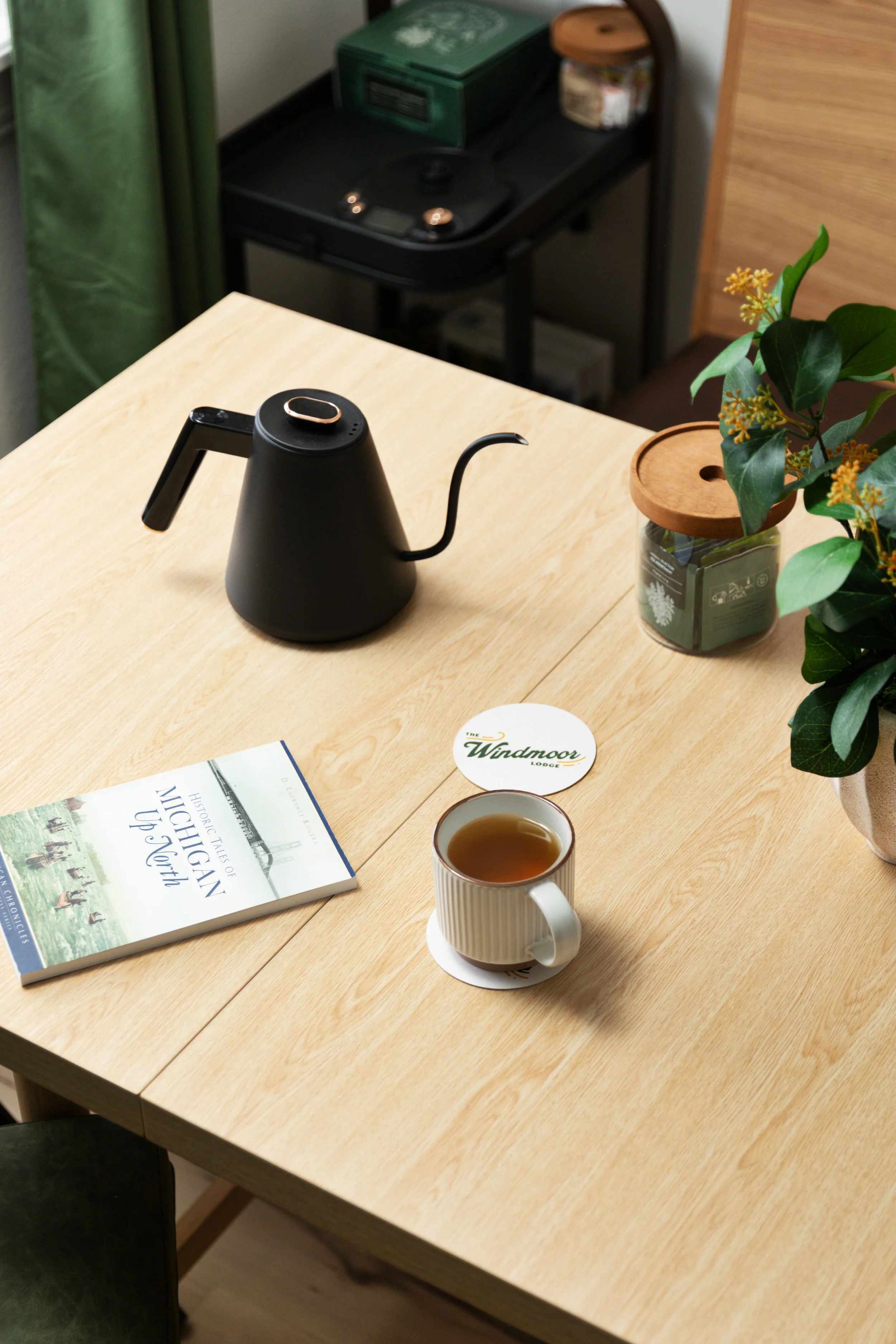 A wooden table with a white cup of tea, a book titled "The Historic Tales of Michigan Up North," a green and white coaster, a potted plant, a black electric kettle, and a sticker that says "The Windmoor Lodge."
