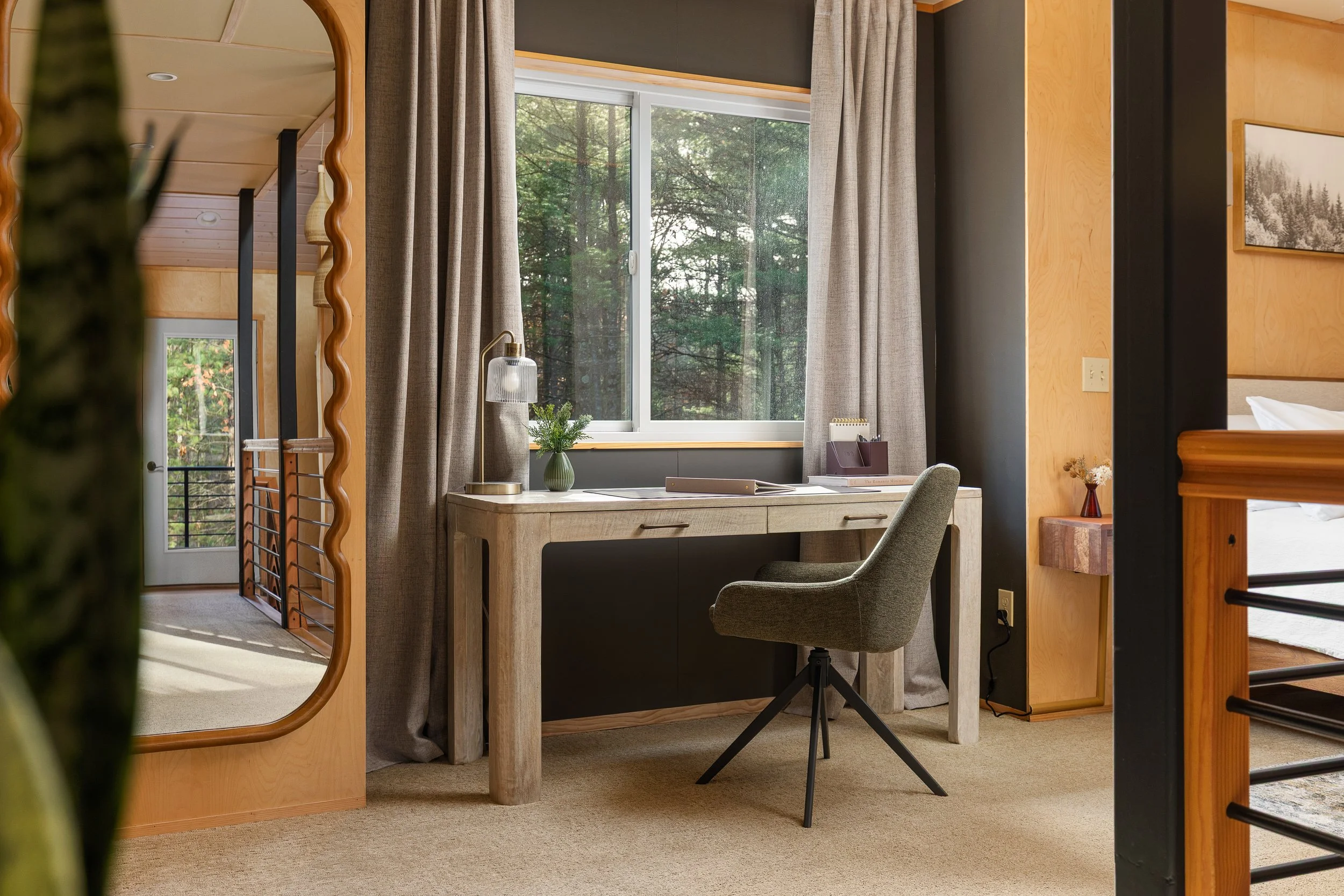 A small home office area with a desk, a chair, a window with green trees outside, and some decor including a flower vase and a desk lamp.