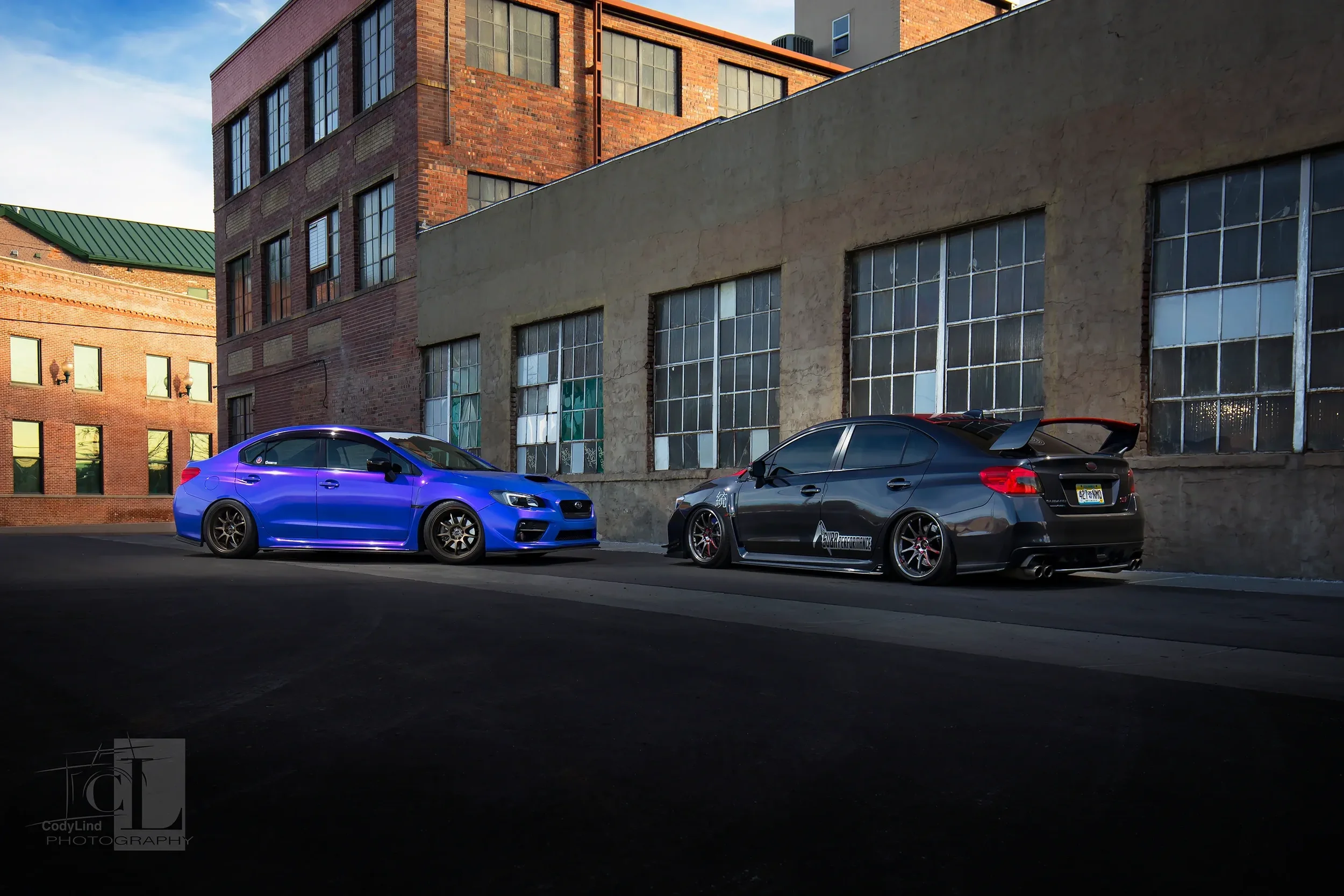 A blue Subaru sports car and a black Subaru sedan with aftermarket modifications parked on a city street against a brick and concrete building with large industrial-style windows.