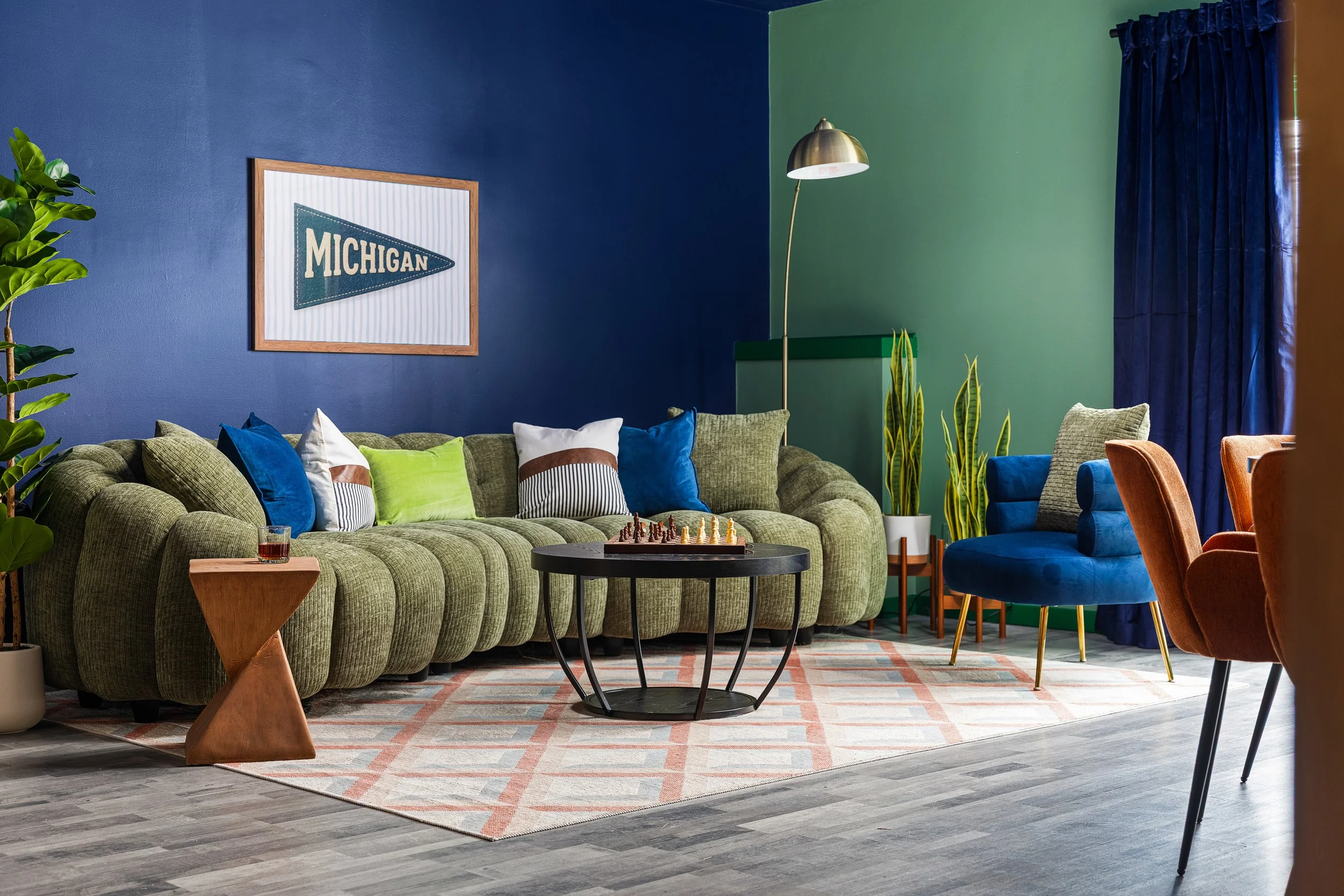 A colorful living room with a green couch, various throw pillows, a black round coffee table with a chess set, a blue armchair, a side table with a drink, plants, a floor lamp, and a framed Michigan pennant on a two-tone wall with dark blue and green
