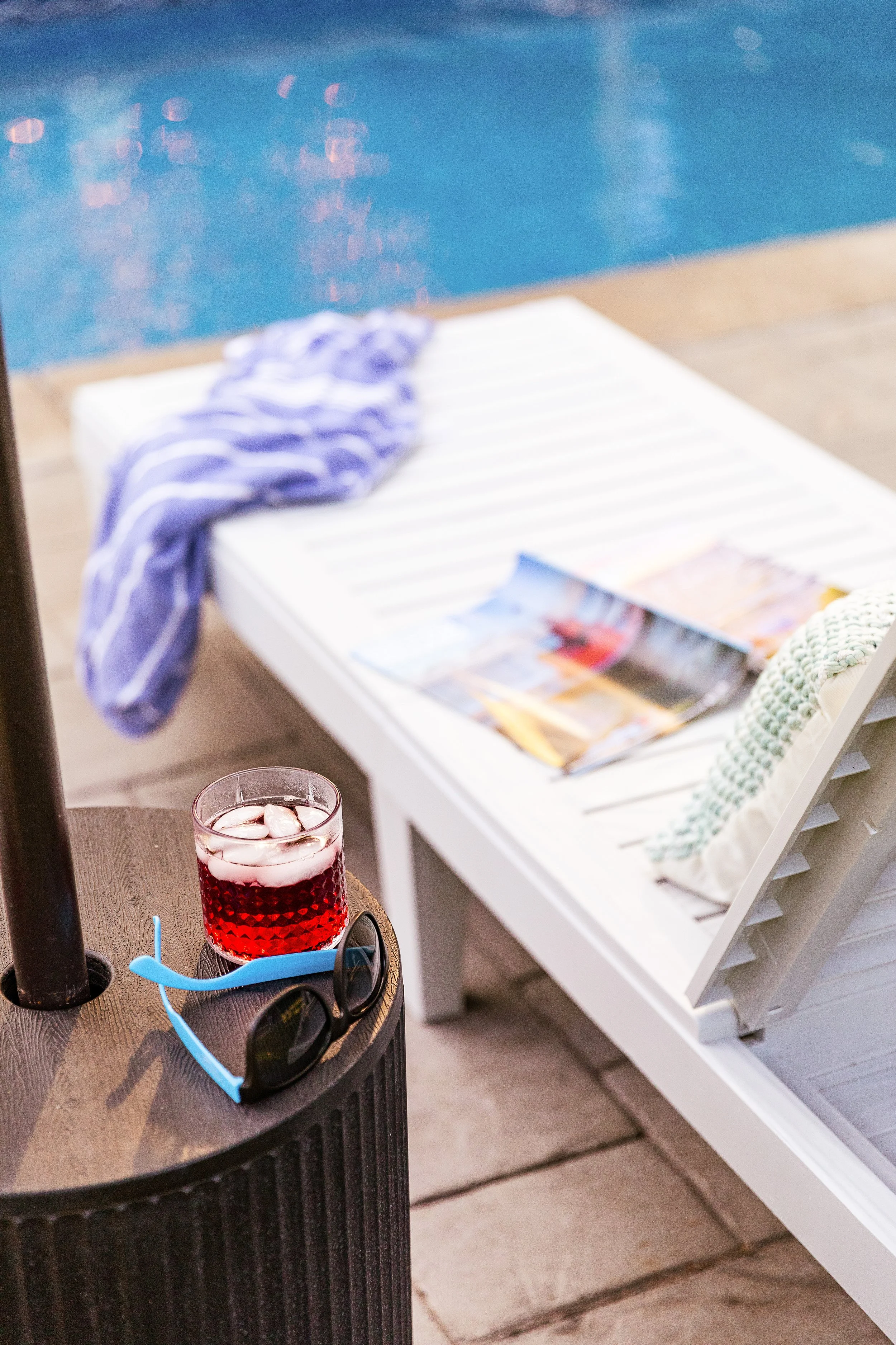 A scene by a poolside featuring a white lounge chair with a magazine and a towel on it, a wooden table with a glass of red drink and sunglasses, and a blue swimming pool in the background.