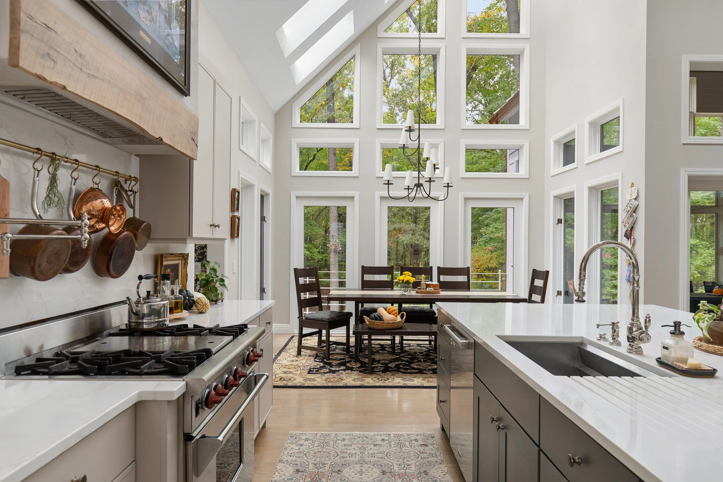 Bright kitchen with white cabinets, stainless steel stove, and a white island with sink. Windows with a view of green trees, a dining table with six chairs, and a chandelier hanging from the ceiling.