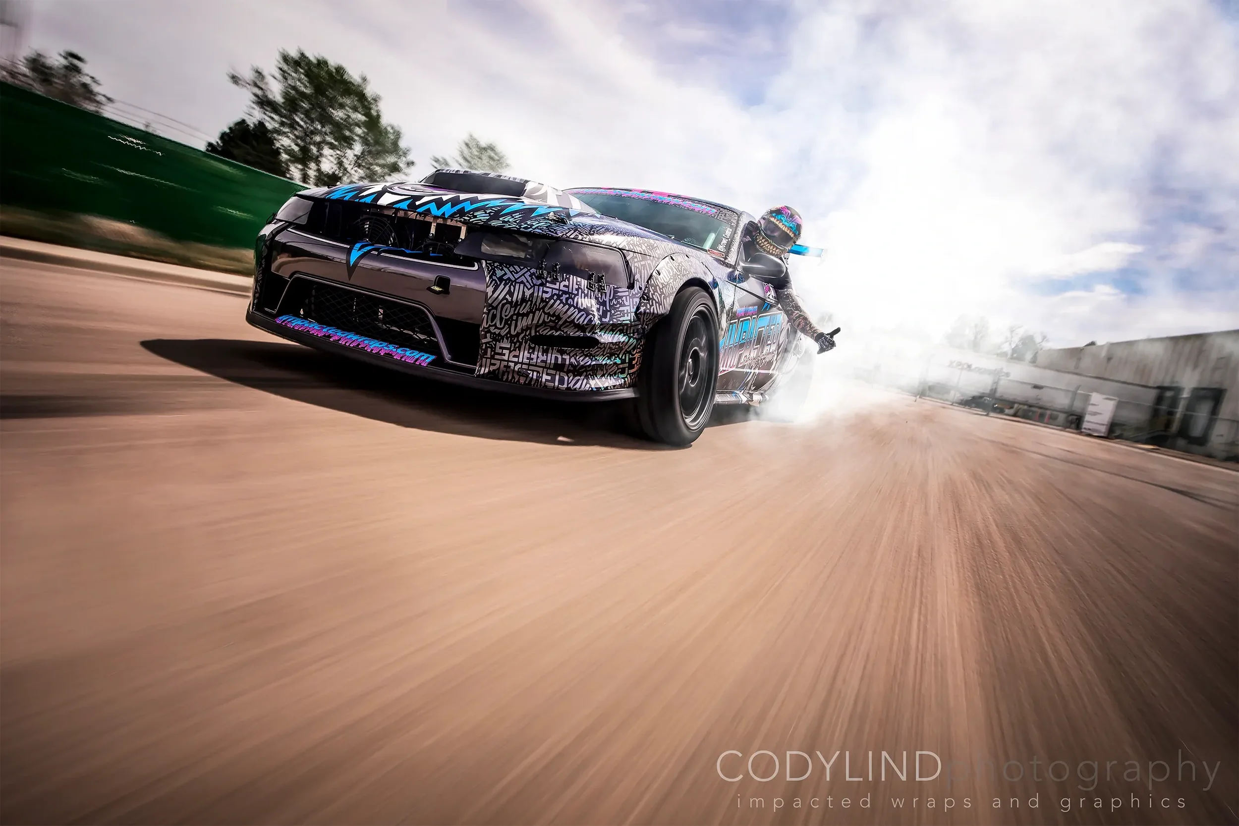 A black race car with colorful graphics speeds along a dirt track, kicking up dust, with the driver extending one hand outside the window, and a cloudy sky overhead.
