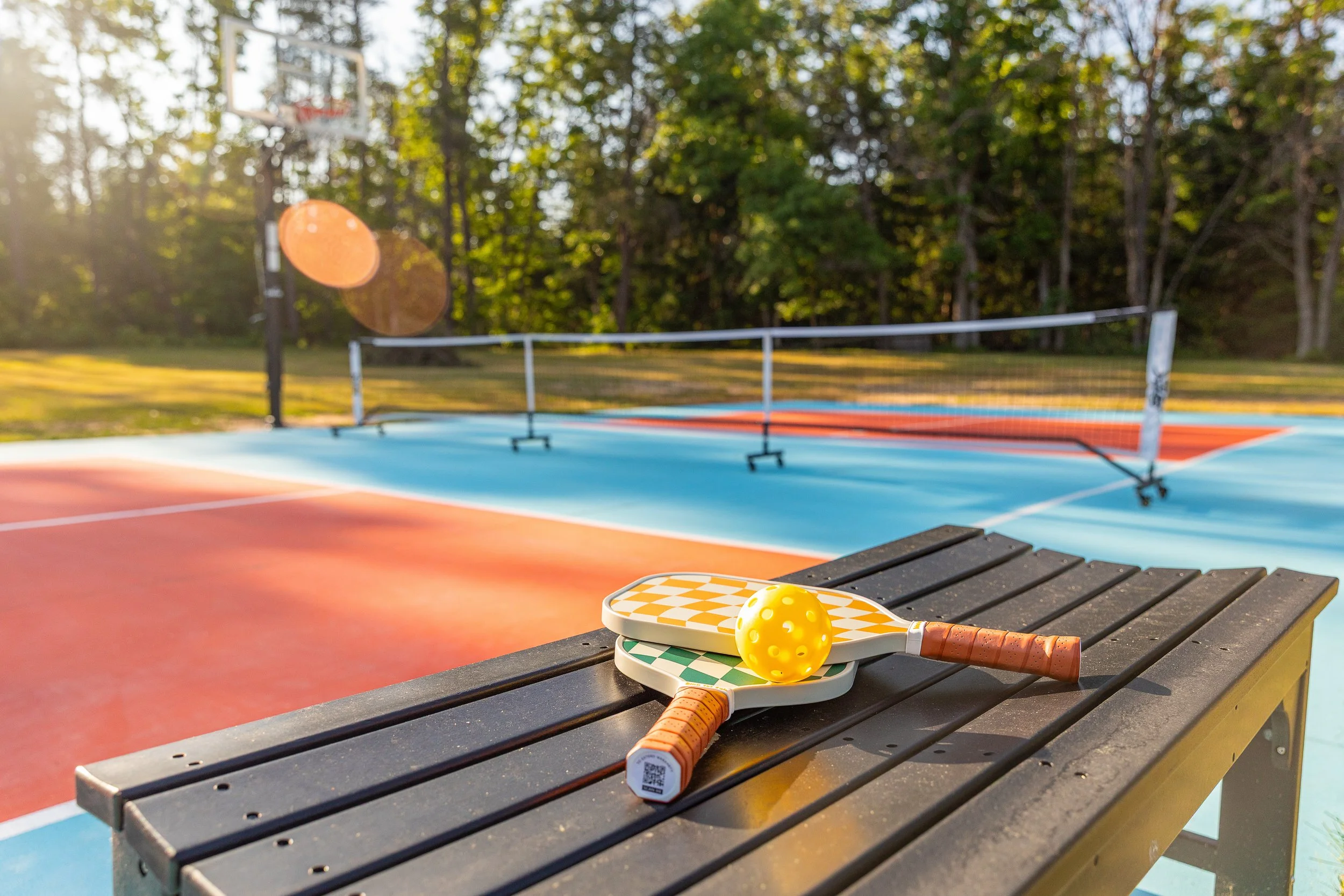 Two pickleball paddles and a yellow pickleball resting on a bench on an outdoor pickleball court during sunset.