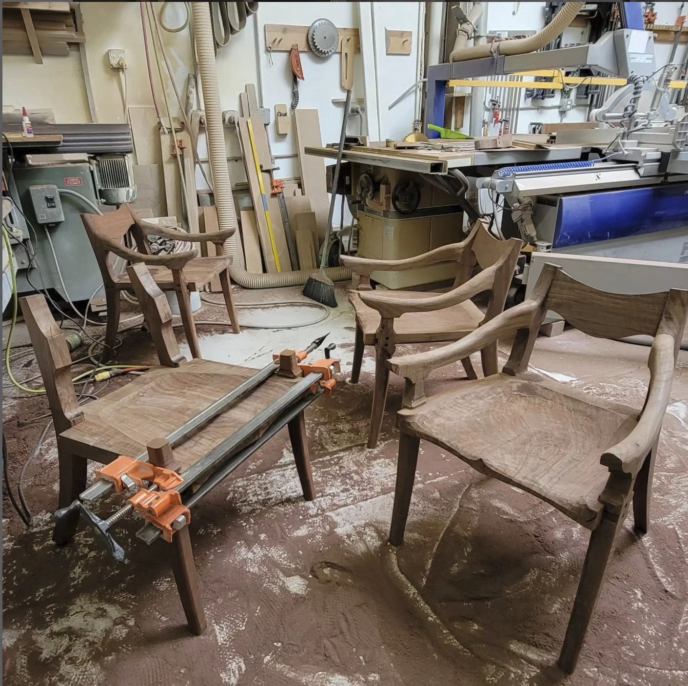 A woodworking workshop with four wooden chairs in progress and various tools, machinery, and wood pieces in the background.
