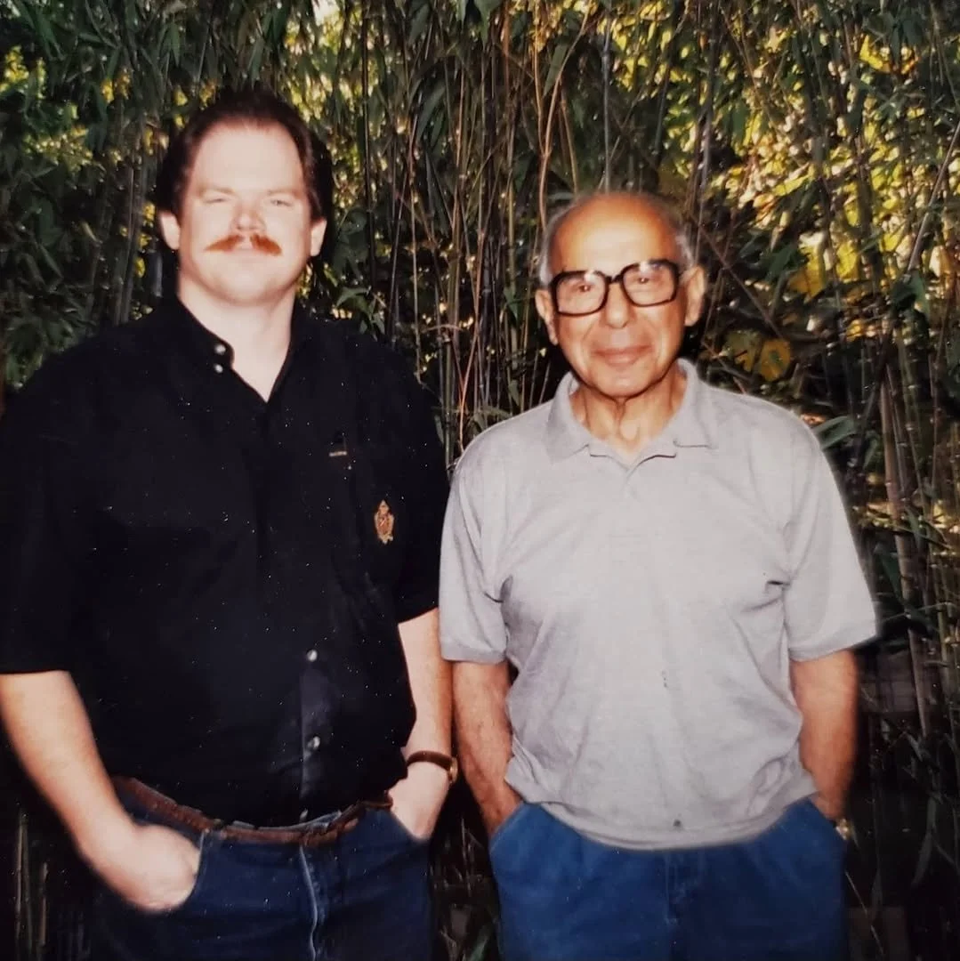 Two men standing side by side outdoors with greenery in the background. One is a younger man with a mustache, wearing a black shirt and jeans. The other is an older man with glasses, wearing a light gray polo shirt and blue jeans.