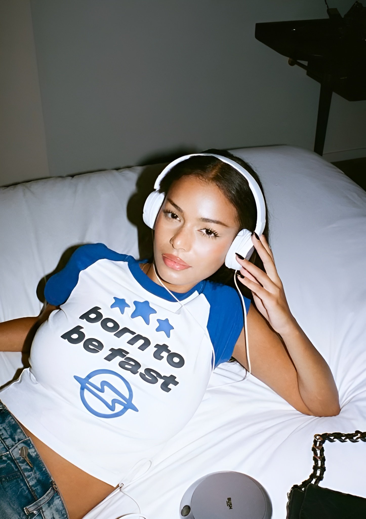 A young woman with long dark hair lying on a bed, wearing headphones and a white and blue t-shirt that says 'Born to be fast' with three blue stars and a lightning bolt symbol.