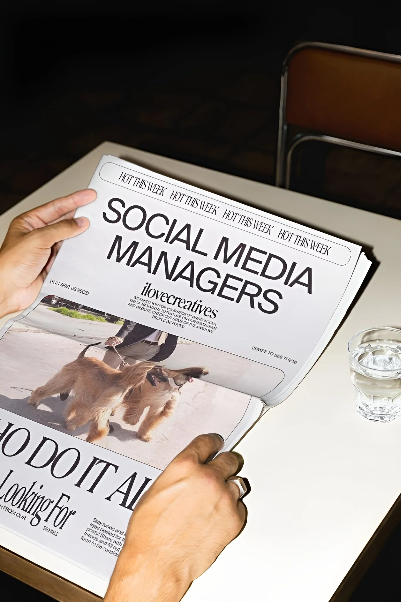Person holding a newspaper about social media managers, sitting at a desk with a glass of water nearby.