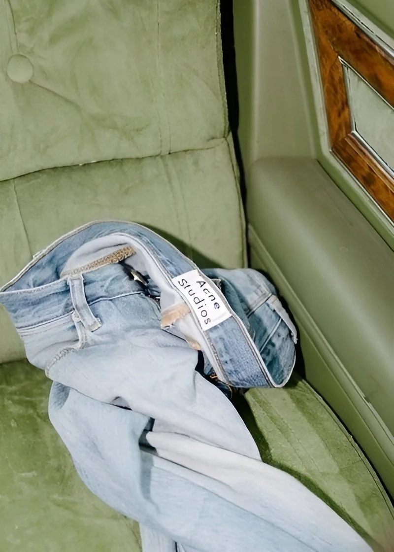 A pair of light-wash jeans with a label that reads 'SOPHIA'S' rest on a green velvet seat near a wall and next to a wood-paneled interior.