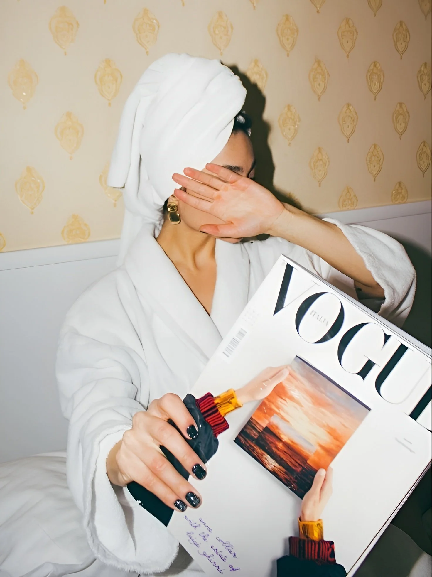 A woman wearing a white bathrobe and a white towel wrapped around her head is holding a Vogue magazine with a sunset photograph on the cover. She is covering her face with her left hand while her right hand is holding the magazine and a small plastic bottle, with a drawing of a hand on it. The woman has dark nail polish and is wearing gold earrings. The background has a patterned wallpaper.
