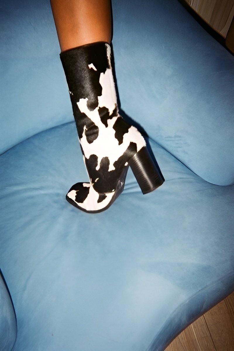 A woman wearing a black and white cow-patterned ankle boot with a chunky heel, sitting on a light blue cushioned chair.