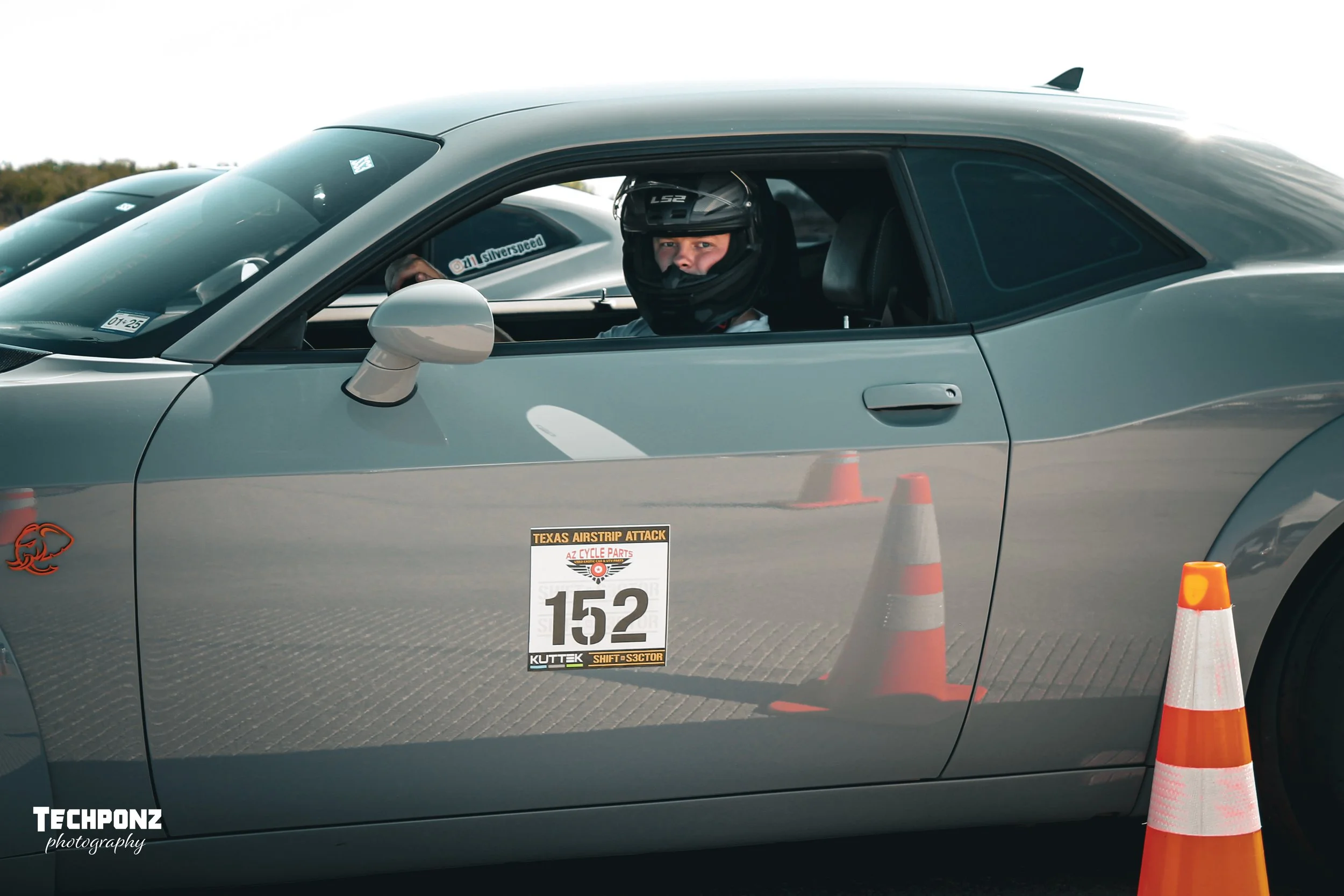 A person in racing gear with a helmet sitting in a silver sports car at a racing event, with traffic cones nearby and a sign displaying the number 152 on the car door.