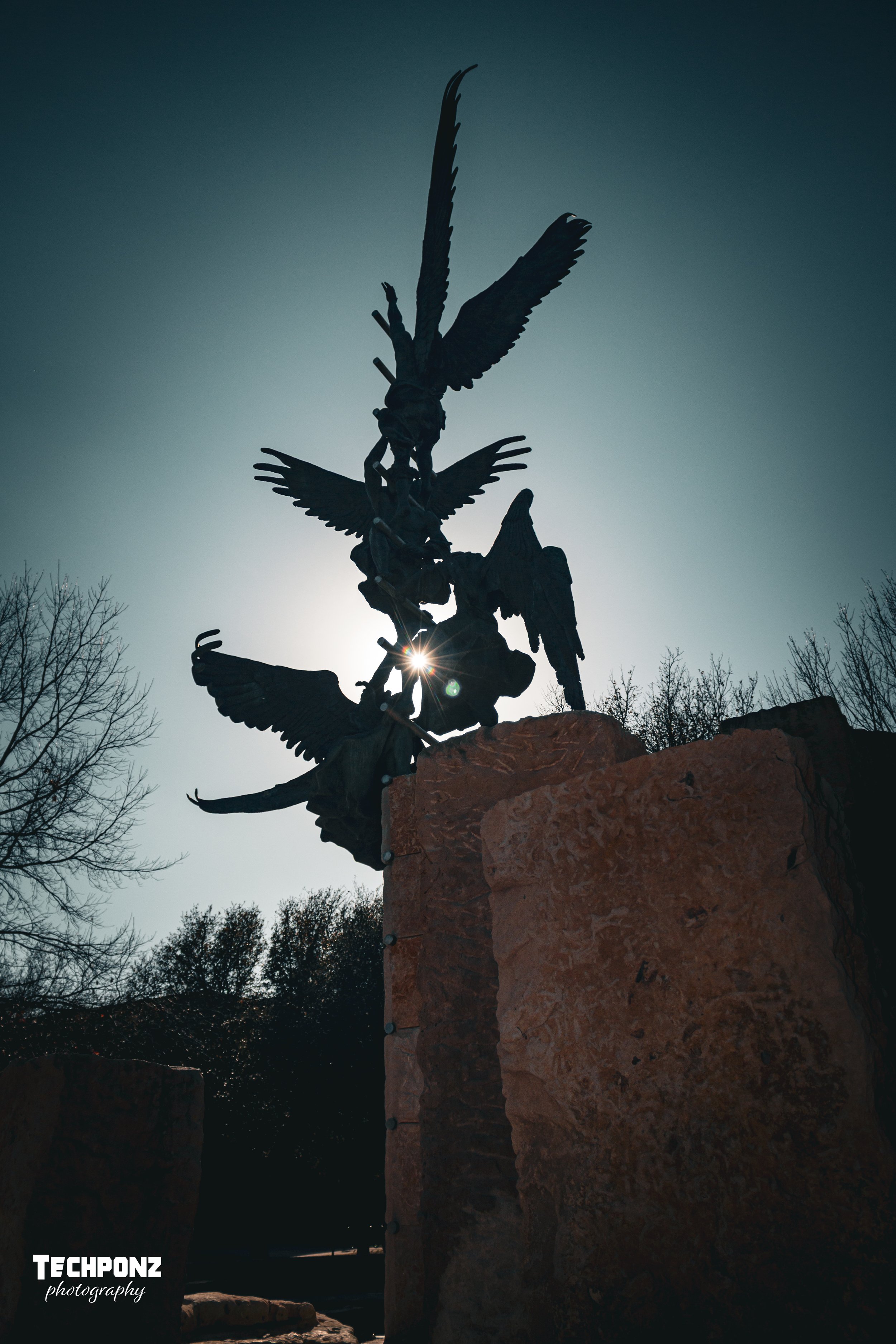 Silhouette of a multi-winged angel statue with a sword, set against the sun, on a stone pedestal, with leafless trees in the background.