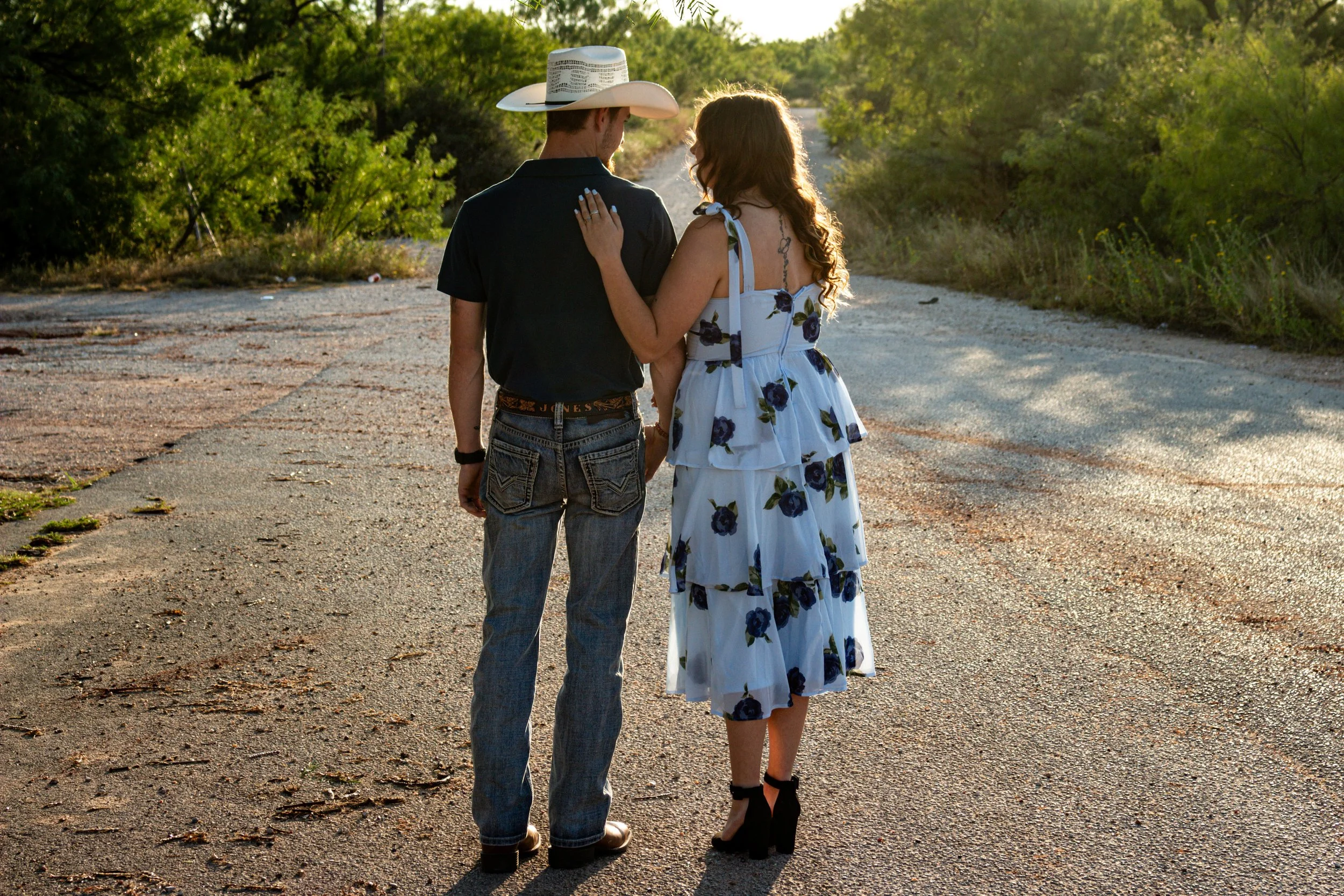 A couple holding hands and walking on a rural, weathered road in a green, wooded area during sunset. The woman is wearing a floral dress and high heels, and the man is wearing jeans, a black shirt, and a cowboy hat.