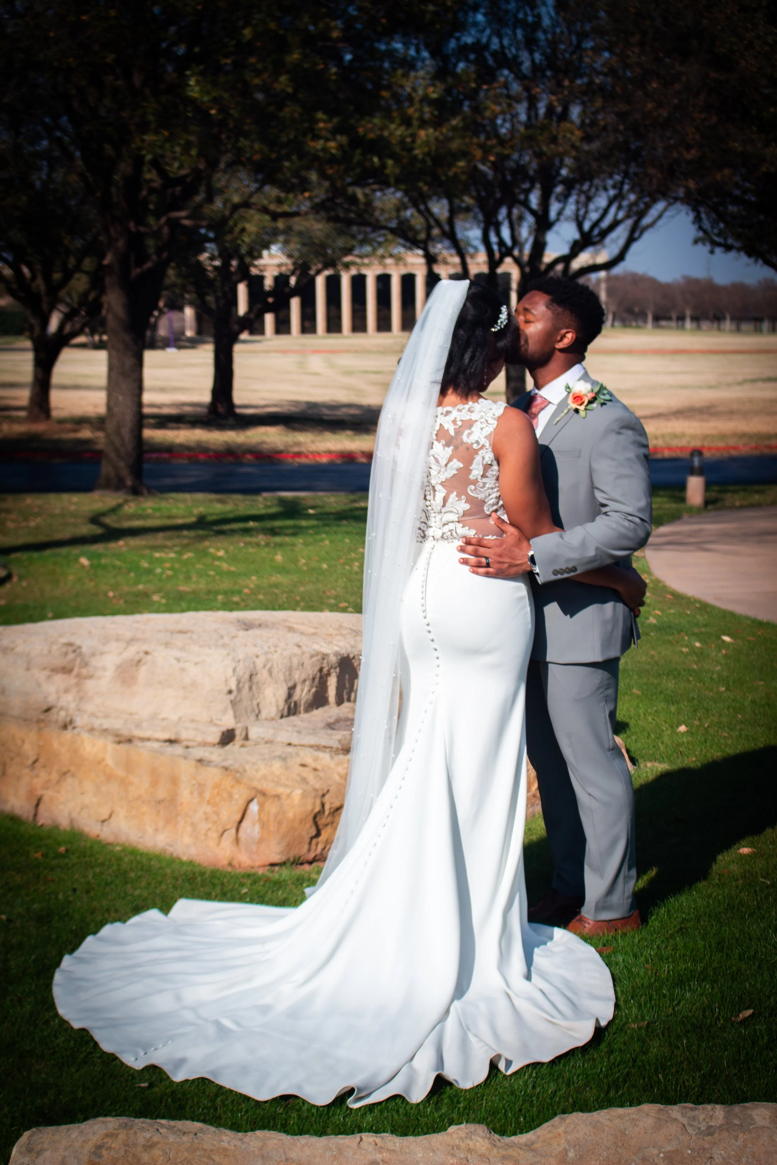 A bride and groom are kissing outdoors on a sunny day, with trees and a classical building in the background.