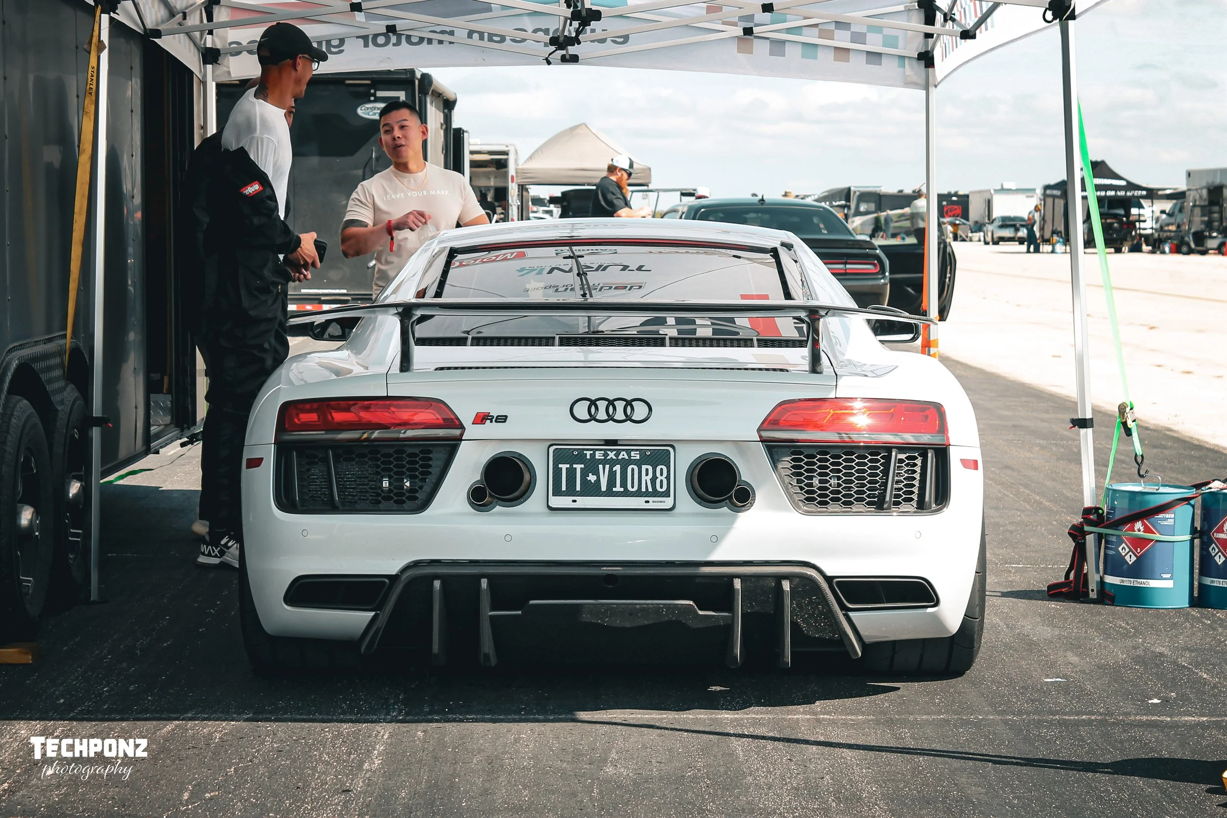 A white Audi R8 race car with Texas license plate, parked under a canopy, with two men standing nearby having a conversation. The car is in a race pit area with trailers and other cars in the background.