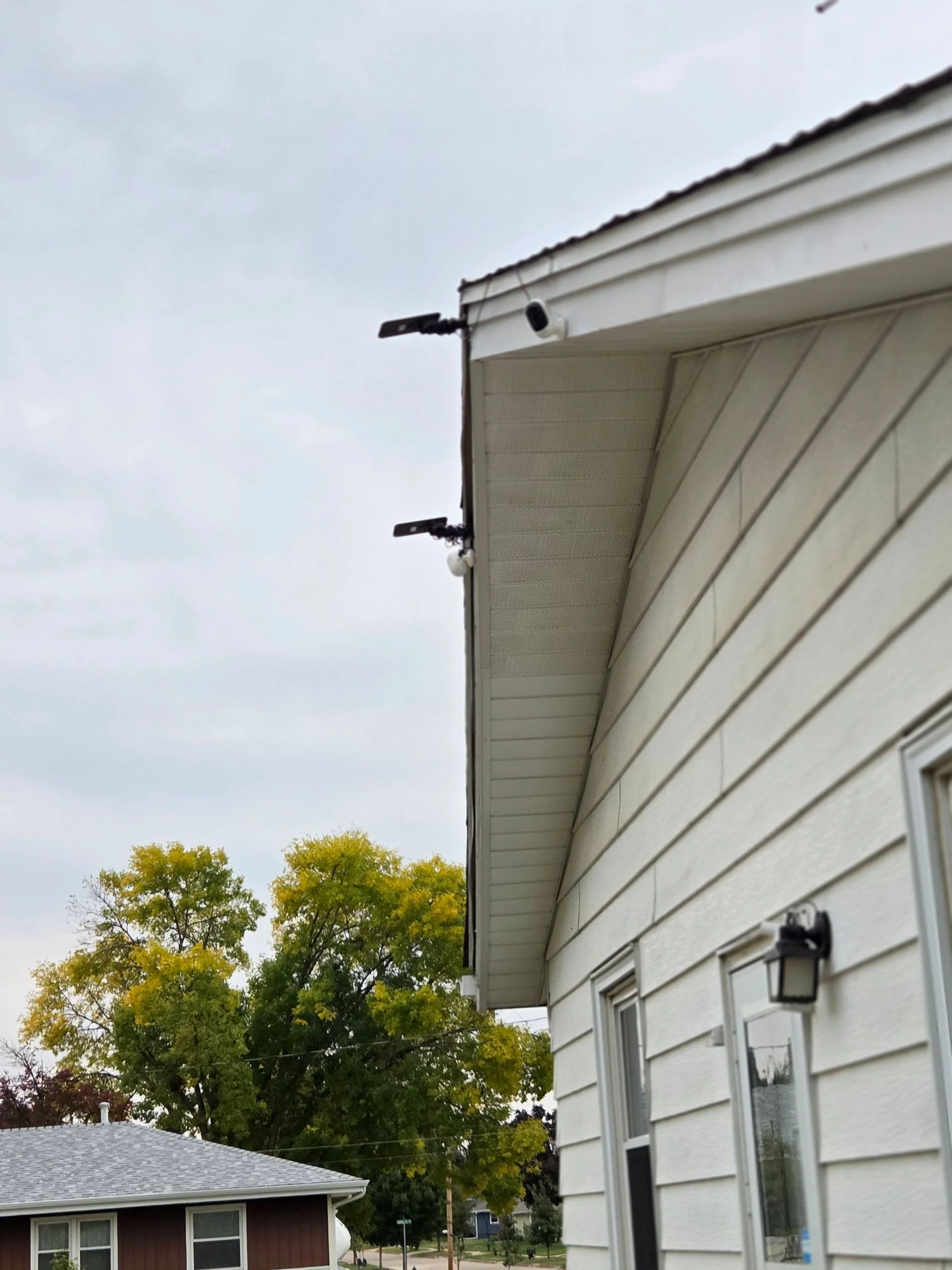 Close-up of the exterior side of a white house with beige siding, showing two outdoor wall-mounted light fixtures and two small cameras attached to the overhang of the roof, and trees in the background.