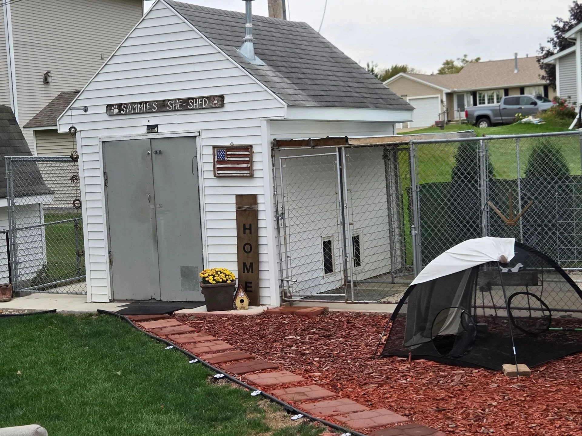 Small white shed with a sign that reads 'Sammies She Shed', decorated with a wooden American flag and a flower pot with yellow flowers outside the door. A black mesh outdoor tent is placed on reddish wood chips in the yard, with a green grass lawn in the foreground and neighboring houses in the background.