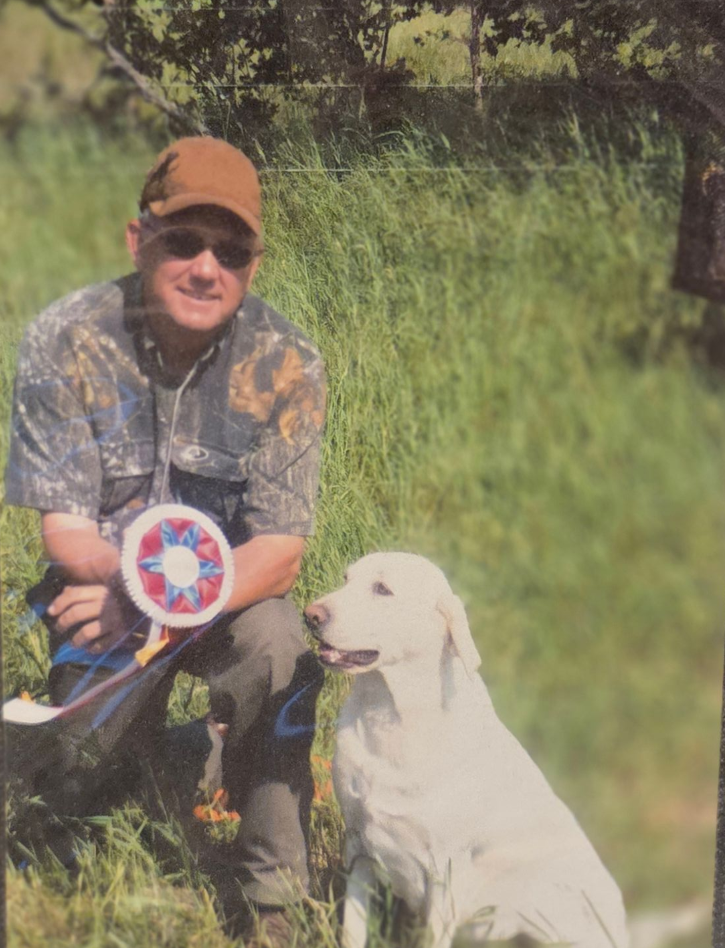 A man crouching in a grassy outdoor area next to a white Labrador retriever. The man is wearing camouflage clothing, sunglasses, and a brown cap. The man is holding a ribbon or award, and the dog is sitting next to him, looking upwards.