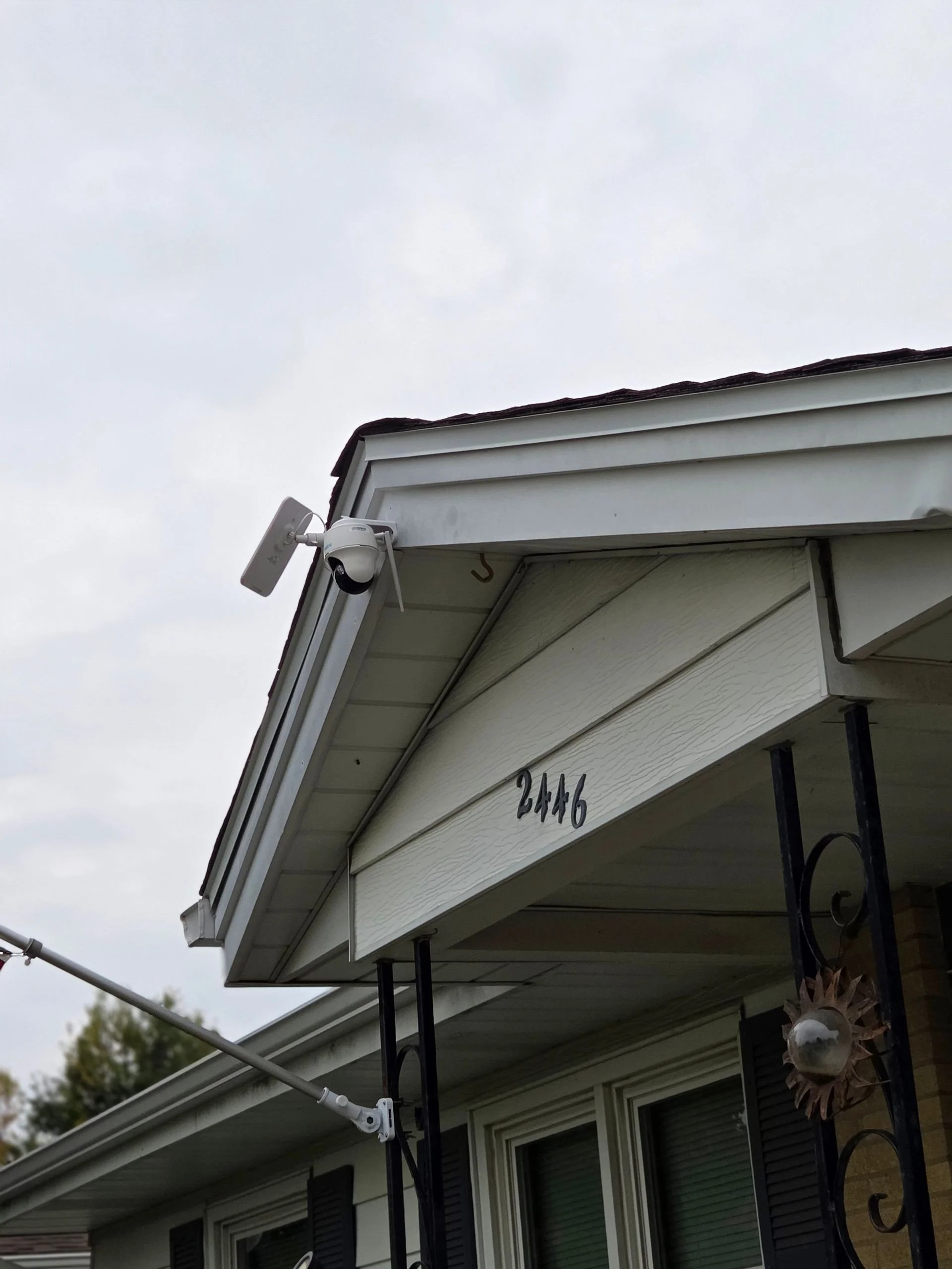 Security camera mounted on the corner of a house with house number 2446 visible, part of a porch with black metal supports and a decorative sun ornament.