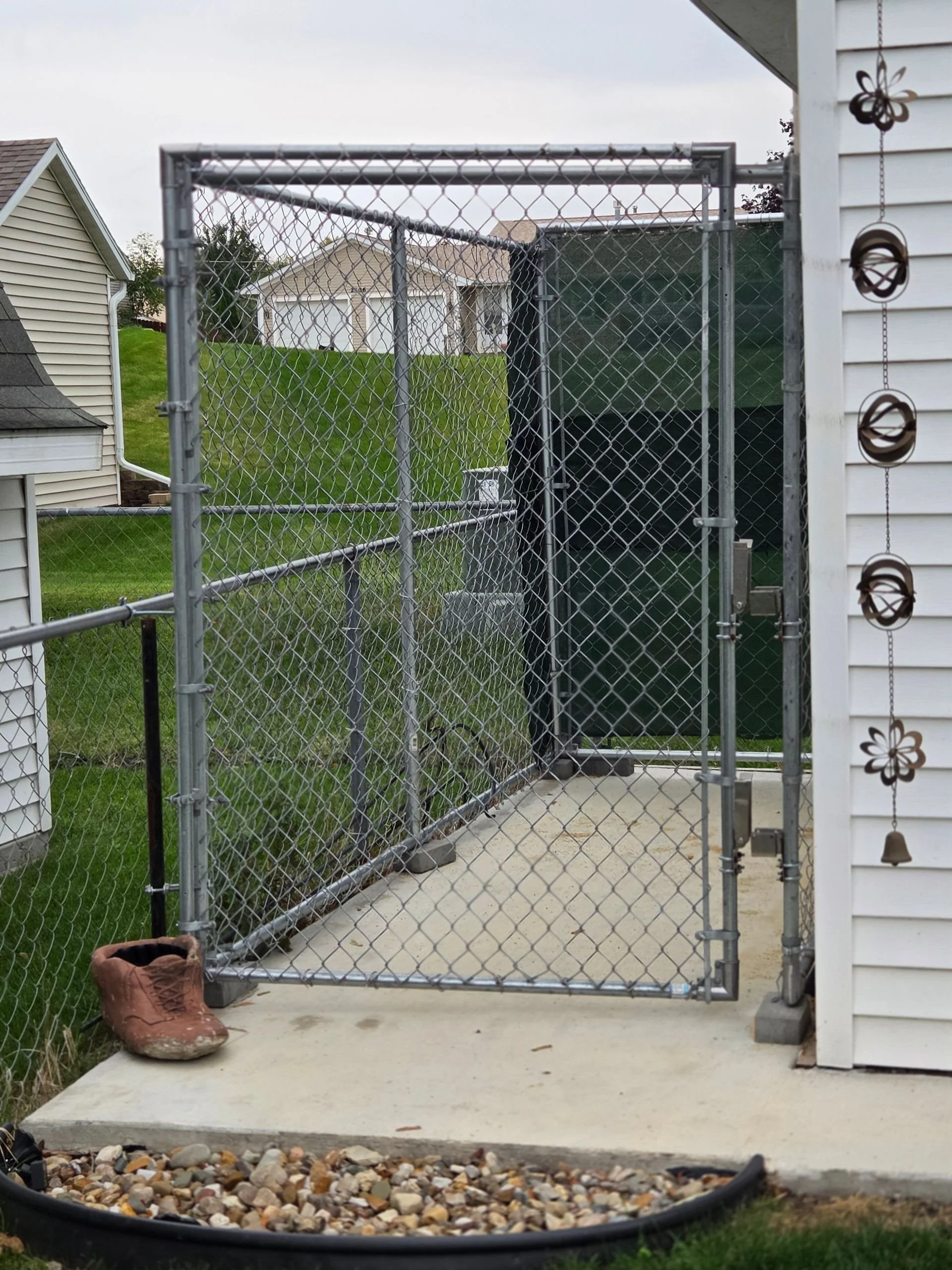 A chain-link fence gate attached to a white house, with a brown boot on the concrete porch and a small border of rocks nearby.