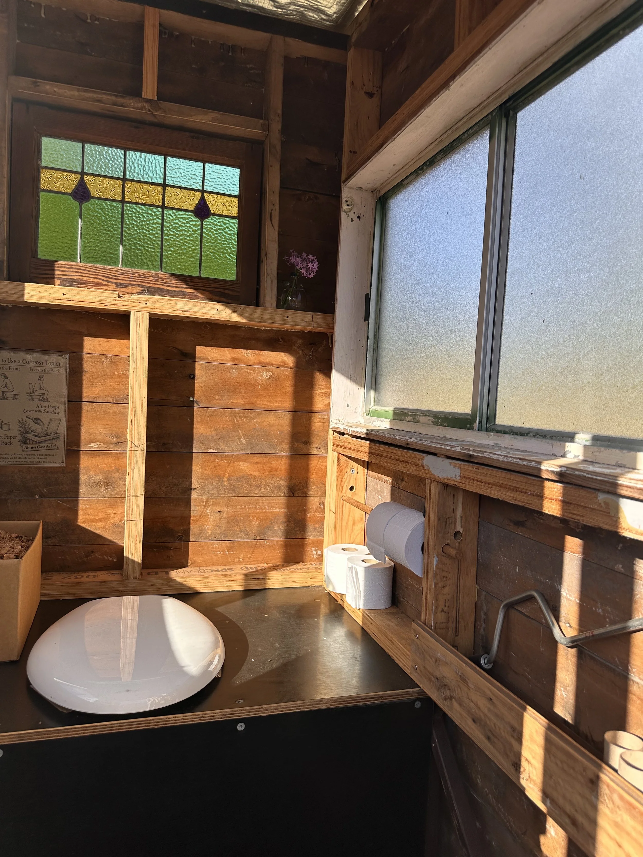 Interior of a small rustic wood-paneled room with stained glass and frosted glass windows, a ceramic sink, and rolls of toilet paper on a wooden shelf.