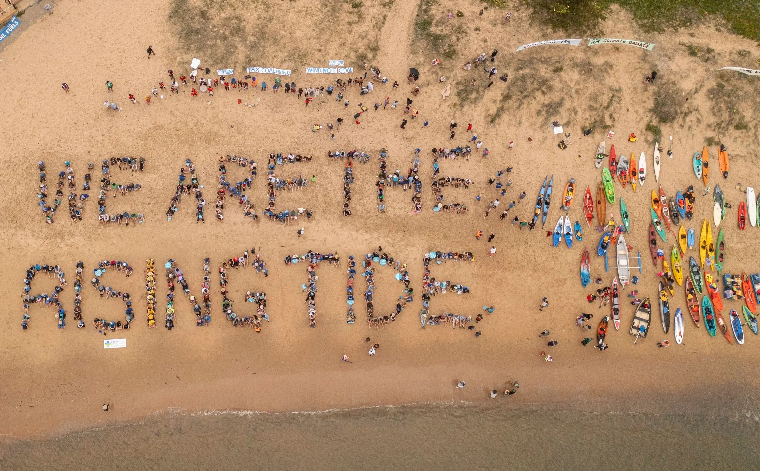Aerial view of a beach where a large group of people are spelling out 'WE ARE THE RISING' with their bodies. To the right, numerous colorful kayaks and boats are lined up on the sand.
