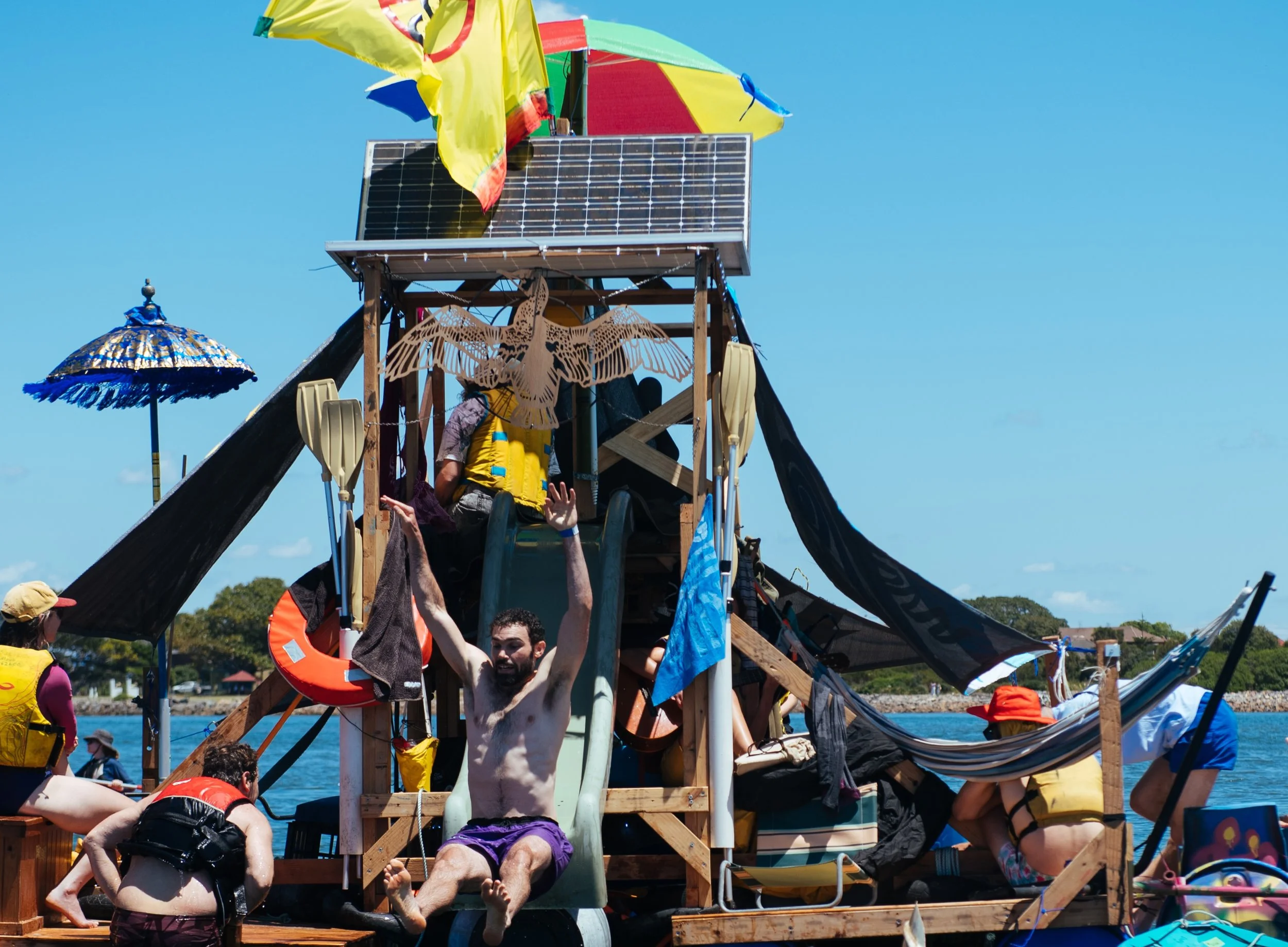 People enjoying a floating water play structure with slides, umbrellas, and decorations on a lake, under a clear blue sky.