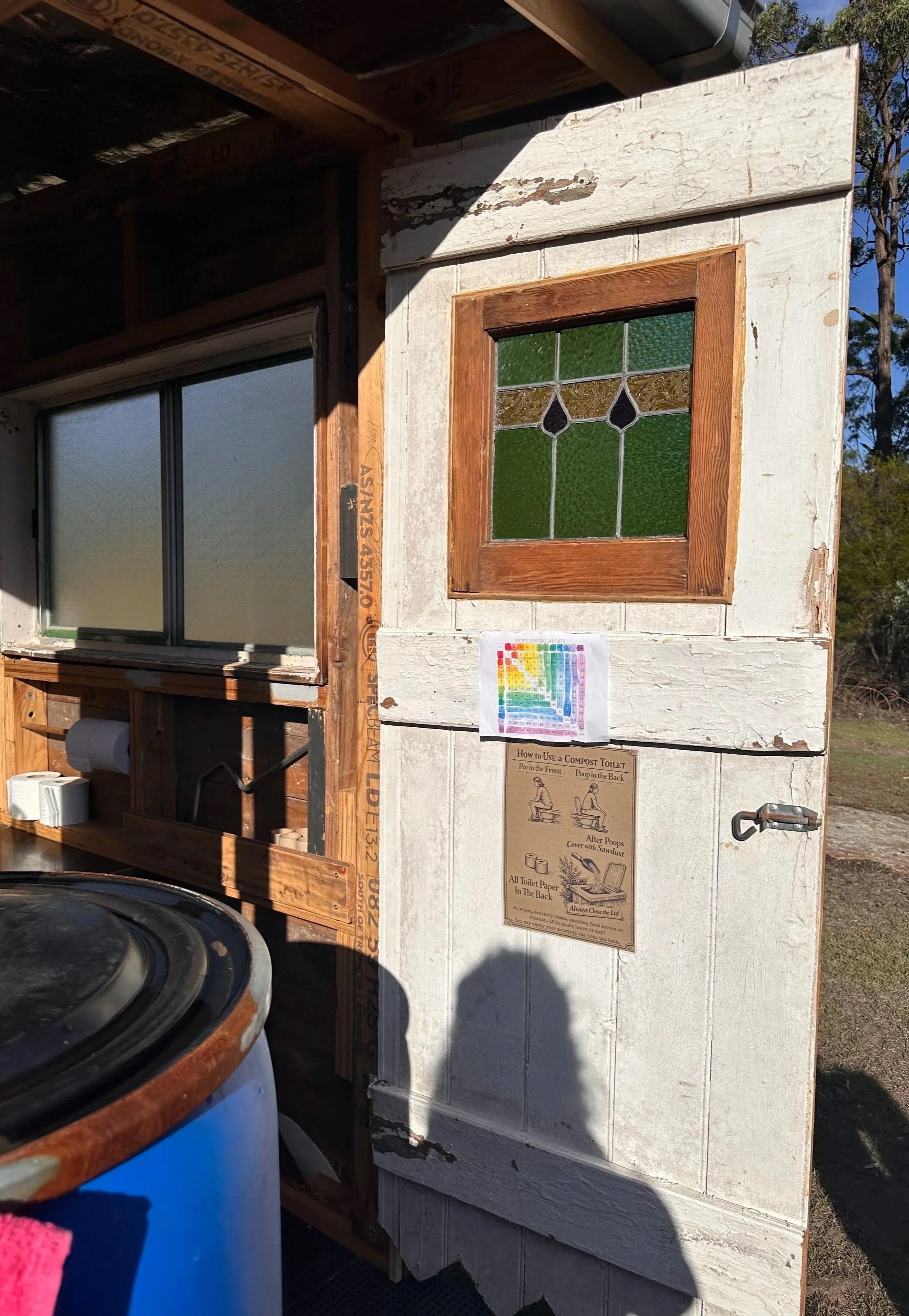 Close-up of a small, weathered building with a stained glass window, a large window, and various signs, including instructions for using a compost toilet.