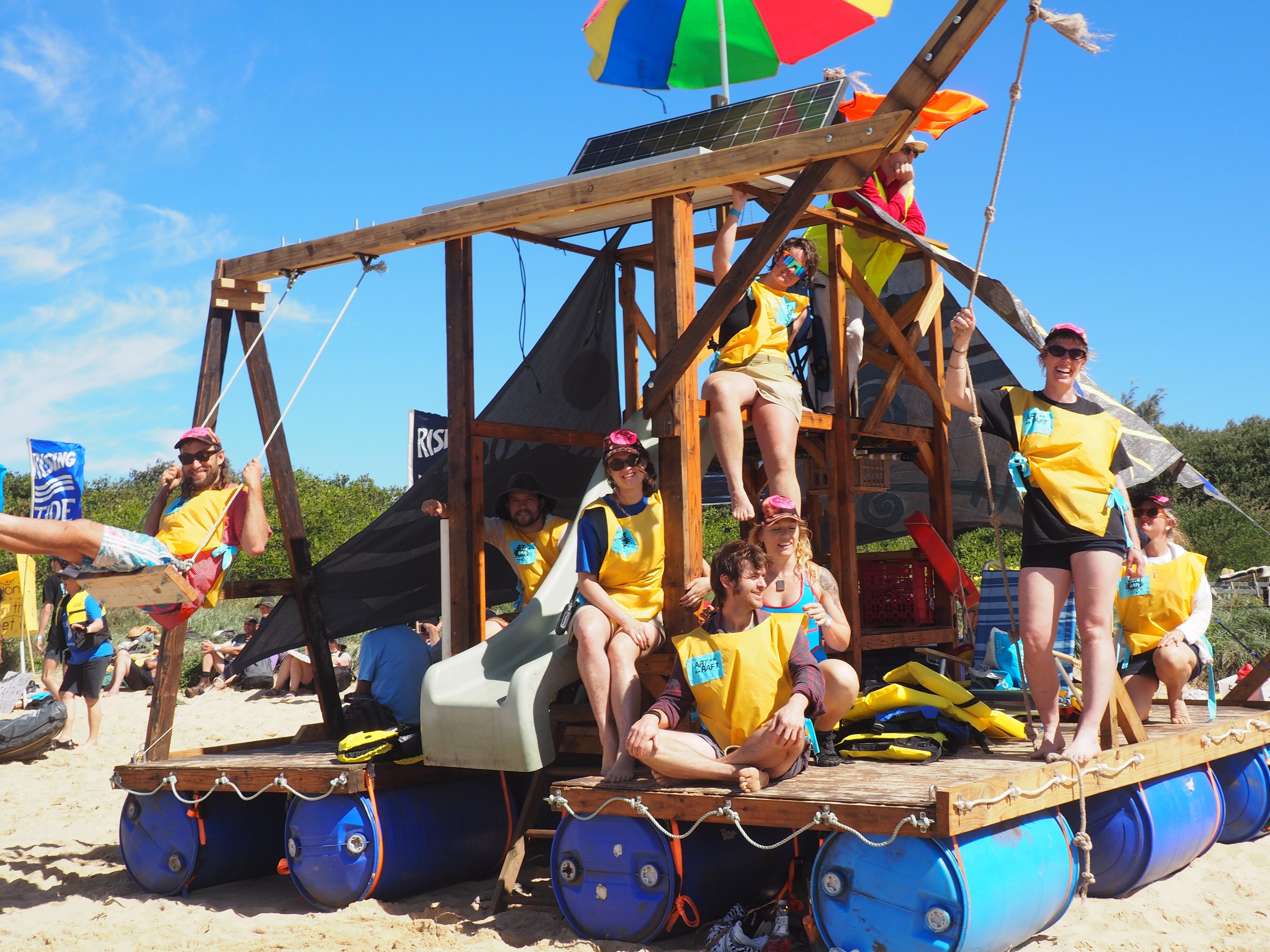 A group of people at the beach on a makeshift float with a wooden structure, swings, and slides, wearing yellow and blue life vests, under a colorful umbrella.