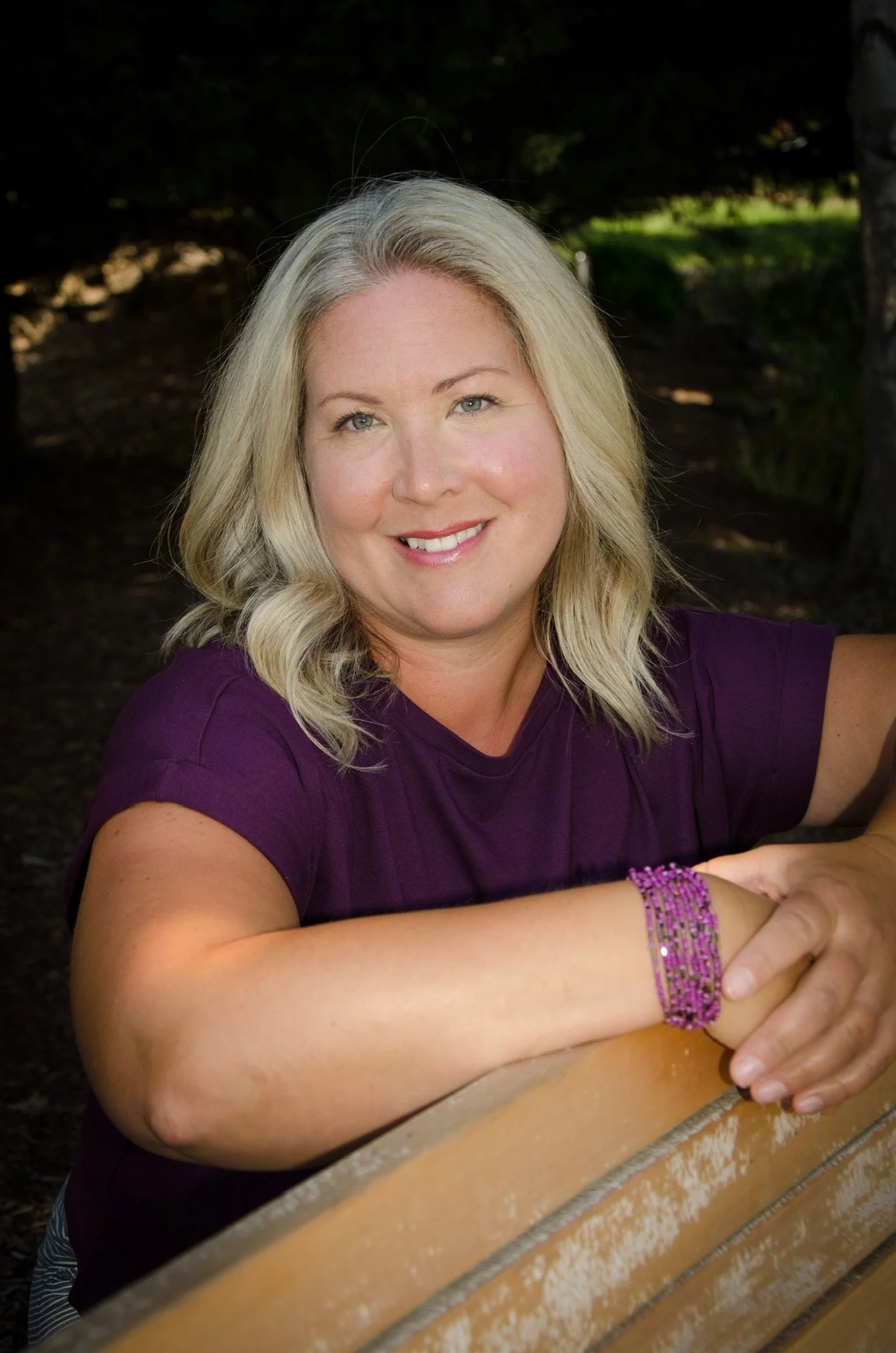 A woman with blonde hair and light skin smiling, wearing a purple shirt and purple beaded bracelet, sitting outdoors on a wooden bench.