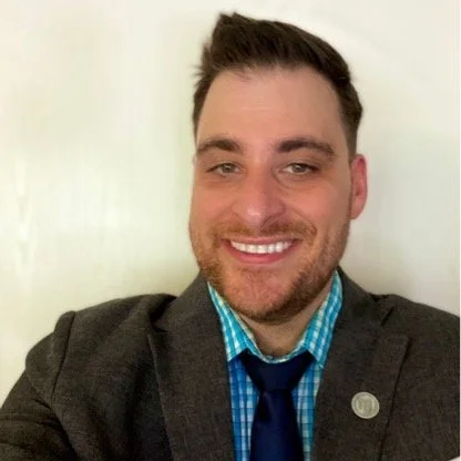 A smiling man with brown hair, wearing a gray blazer, blue checkered shirt, and a dark blue tie, in front of a plain light-colored background.