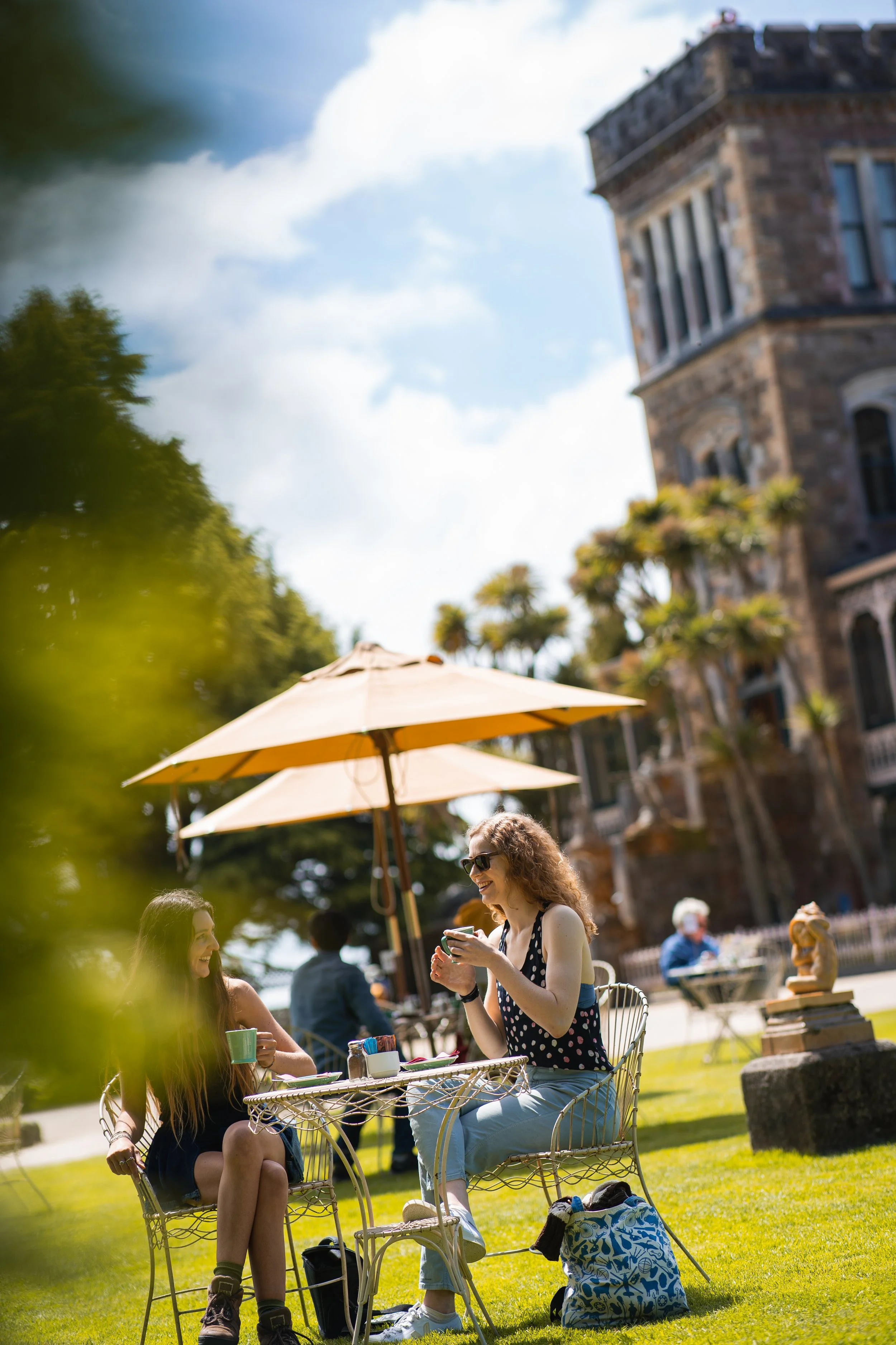 Two women sitting at a metal table with a yellow umbrella in a park, smiling and talking, with a historic building in the background.