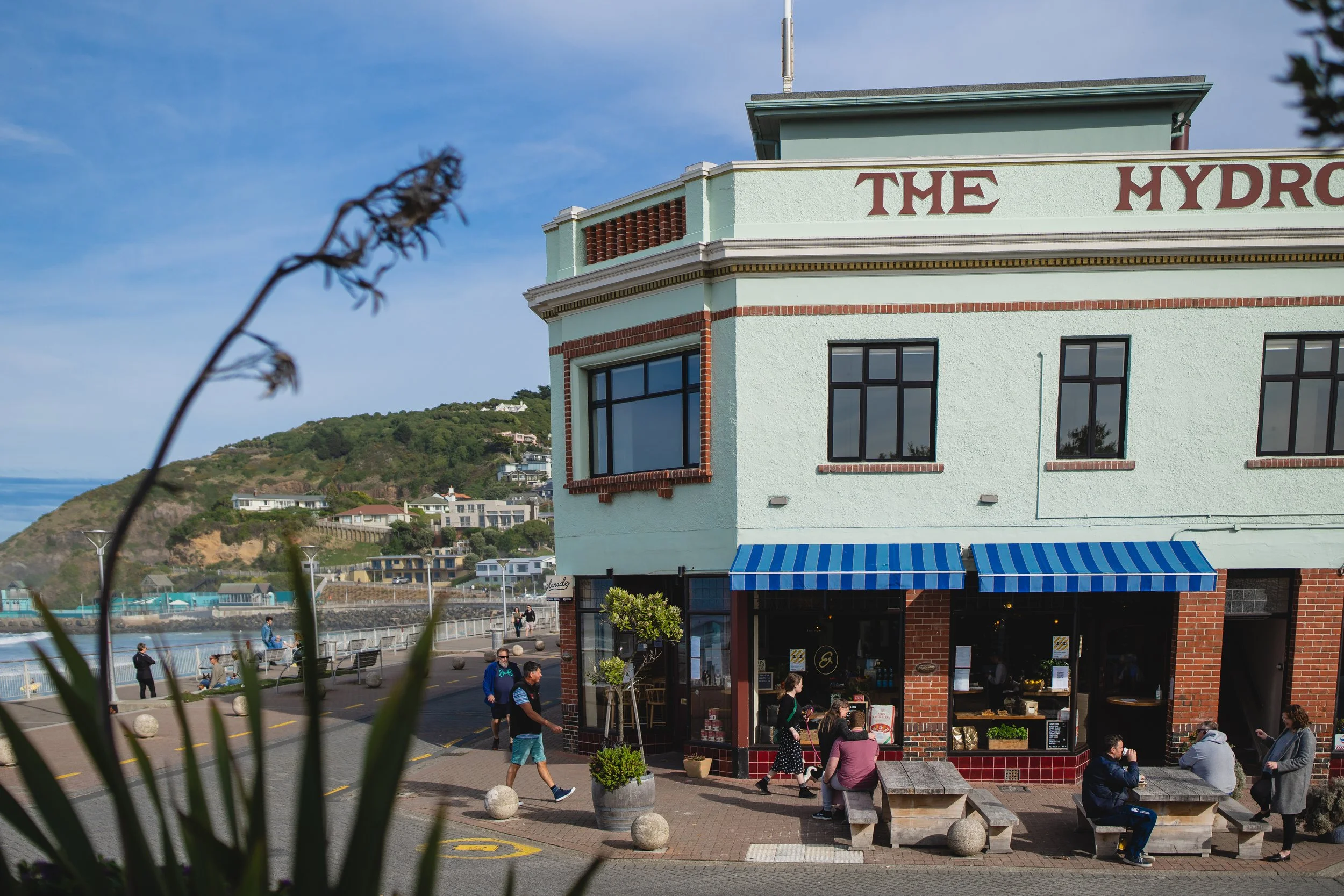 A building by the waterfront with a sign reading 'THE HYDRO' on its upper part, blue and white striped awnings over the entrances, and people sitting and walking outside