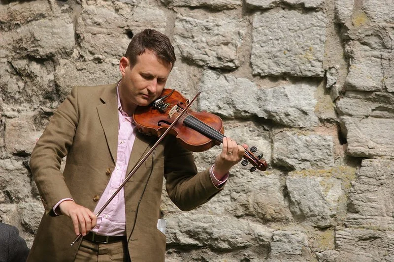 A man wearing a brown suit playing a violin outdoors against a stone wall.
