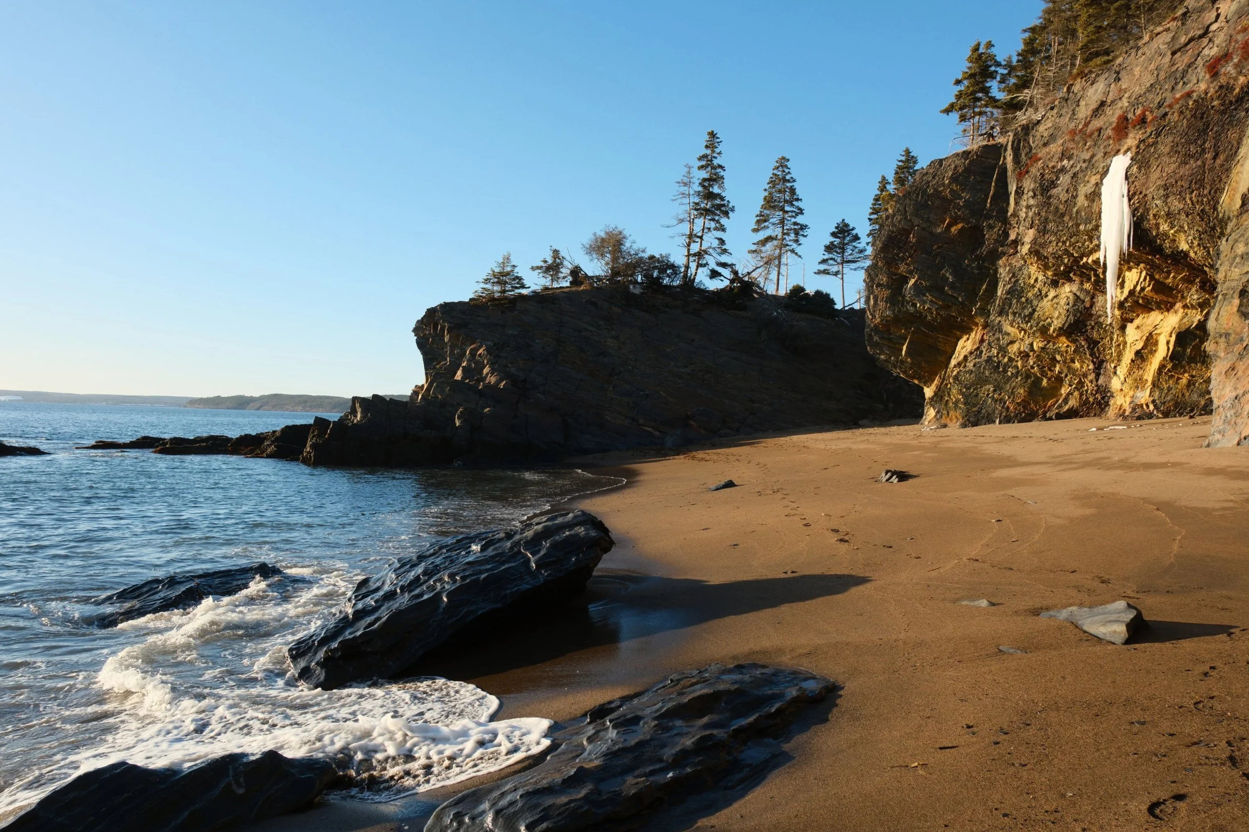 A sandy beach with large rocks near the shoreline, tall cliffs with trees in the background, and calm water under a clear blue sky.