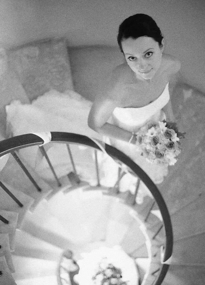 A black and white photo of a woman in a wedding dress, holding a bouquet, standing at the top of a spiral staircase and looking up at the camera.