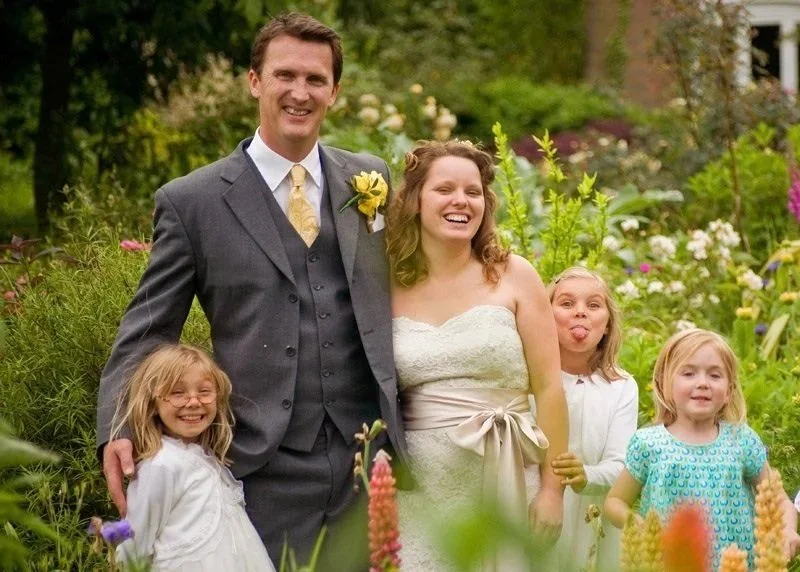 A smiling couple in wedding attire stands with three young girls in a garden filled with colorful flowers.