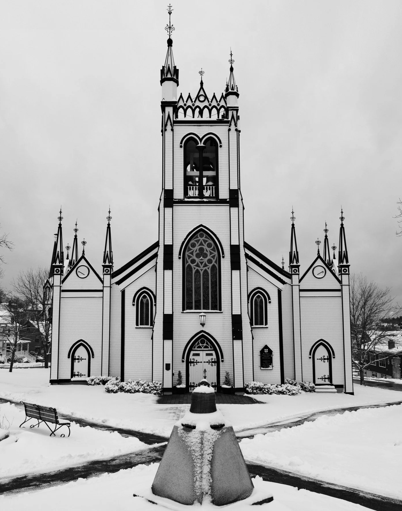 A black and white photo of a church with Gothic architecture, featuring pointed arches, spires, and stained glass windows. The church is situated in a snowy landscape with leafless trees around it.