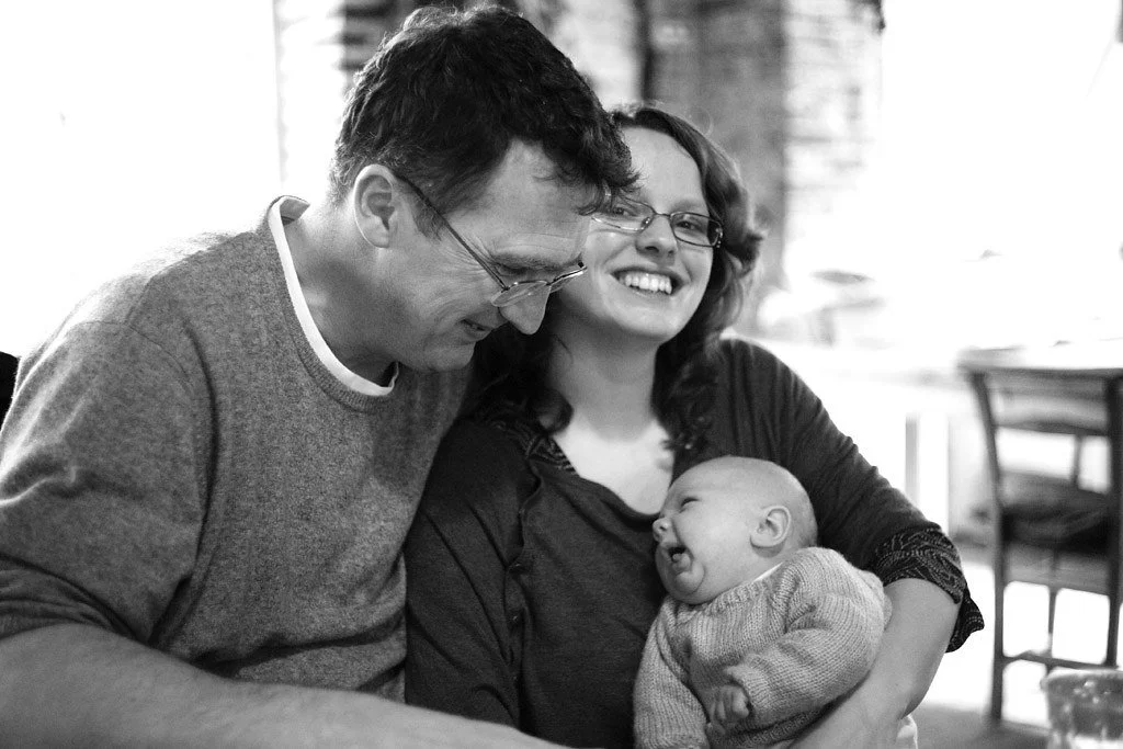A family of three, with a man, woman, and baby, sitting together and smiling. The woman is holding the baby, who is laughing, and the man is leaning in close.