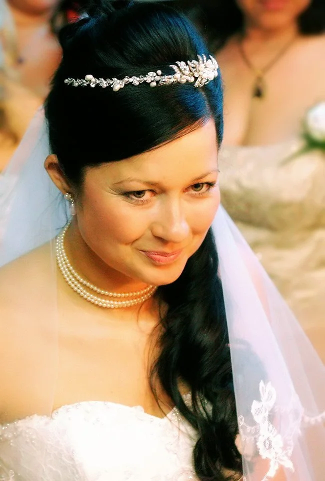 Close-up of a bride with dark hair styled in loose waves, wearing a pearl necklace, pearl earrings, and a tiara, on her wedding day.